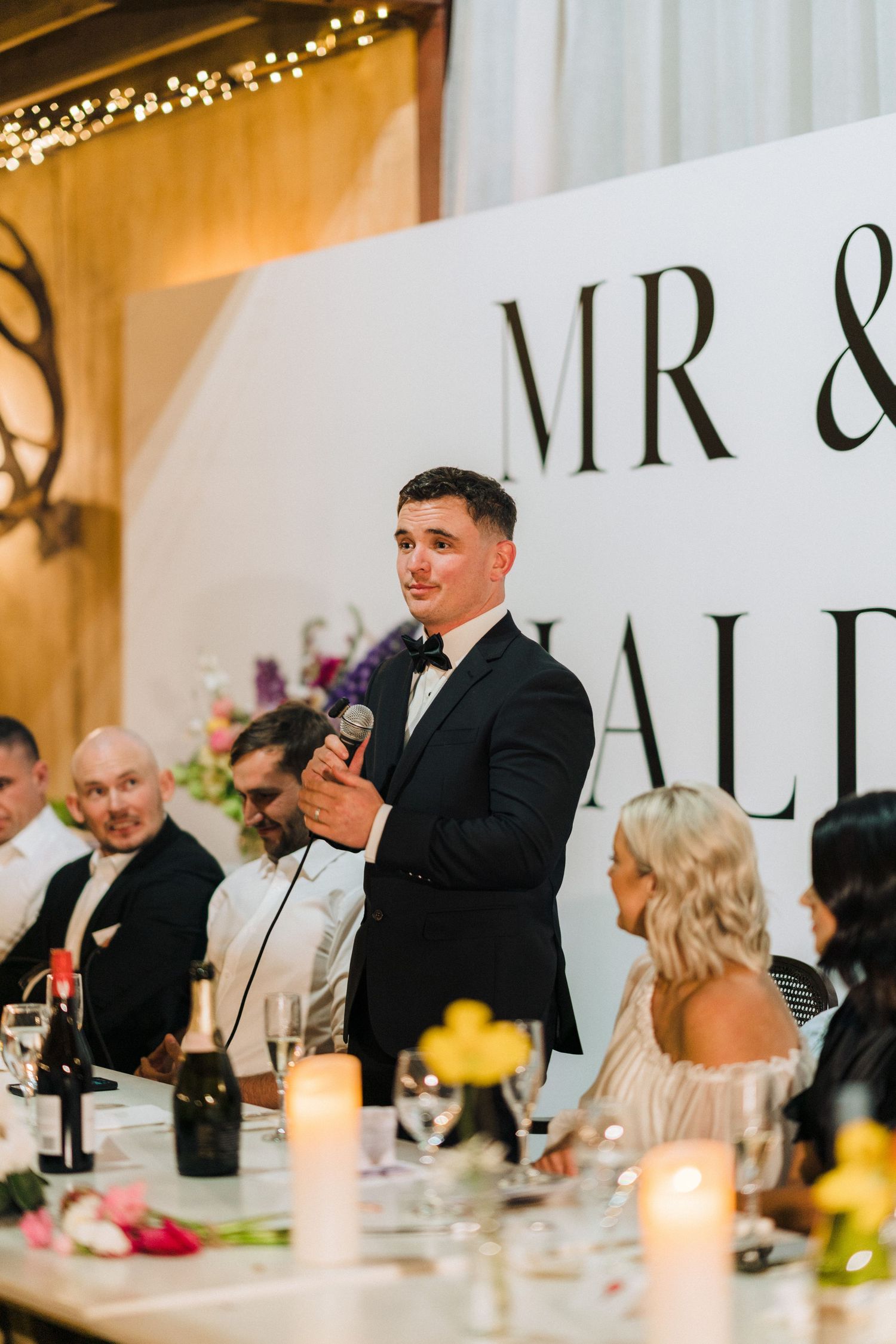 Wedding reception speech at elegant venue with 'MR &' signage, string lights, and formal table settings.