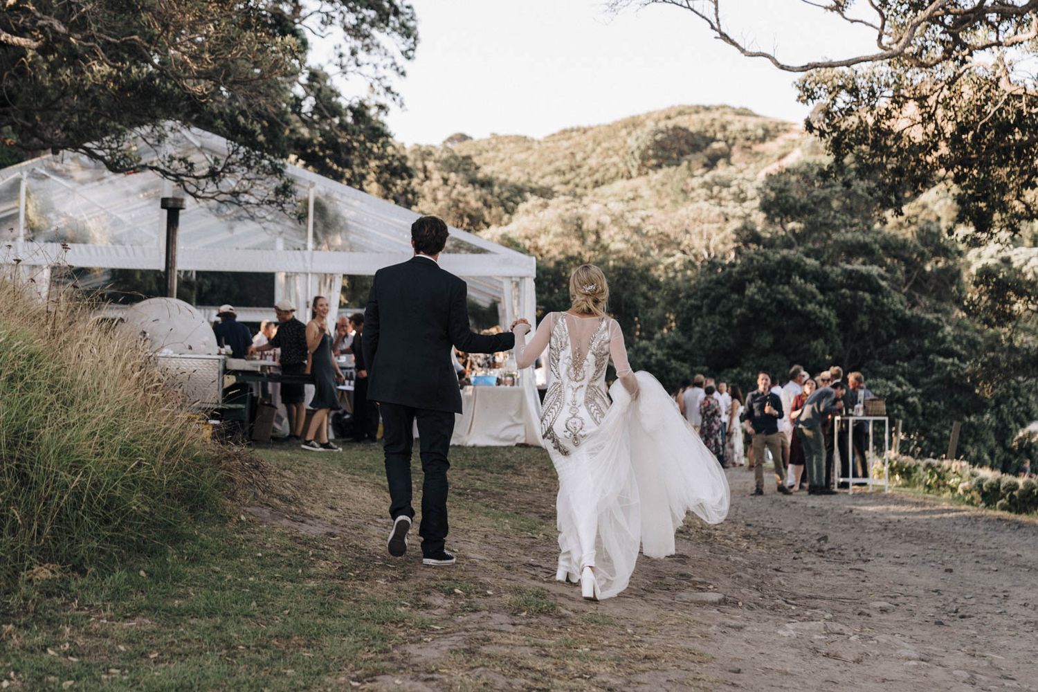 A couple walks along a dirt path towards an outdoor wedding reception venue set against rolling hills.