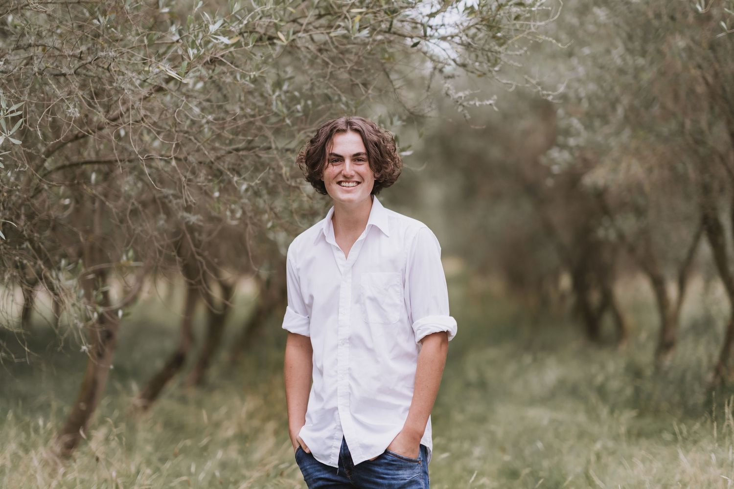 A person in a white button-down shirt and jeans stands casually in a sun-dappled olive grove with soft lighting.