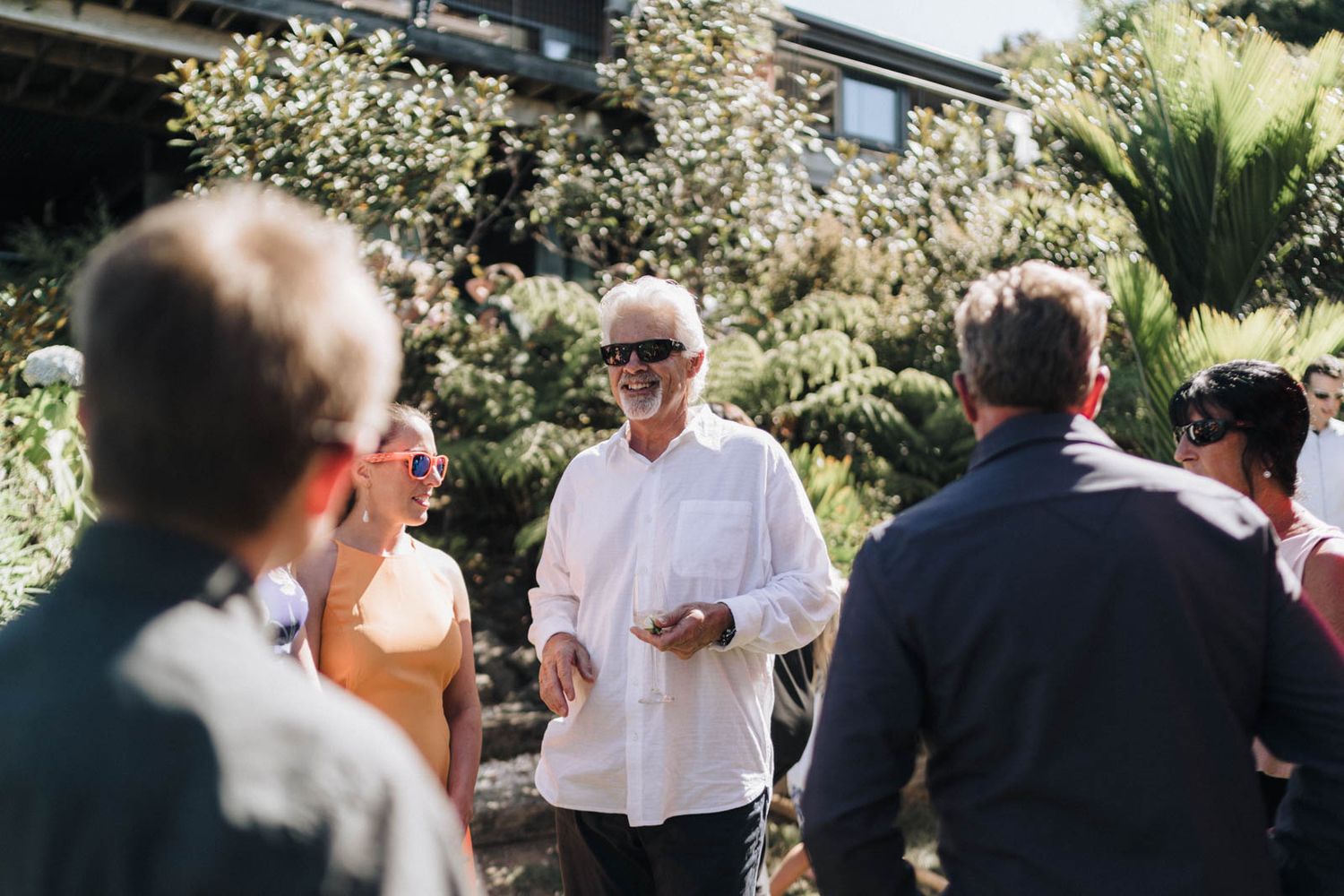Group of people gathered outdoors in a sunny garden setting with lush greenery and flowering plants in the background.