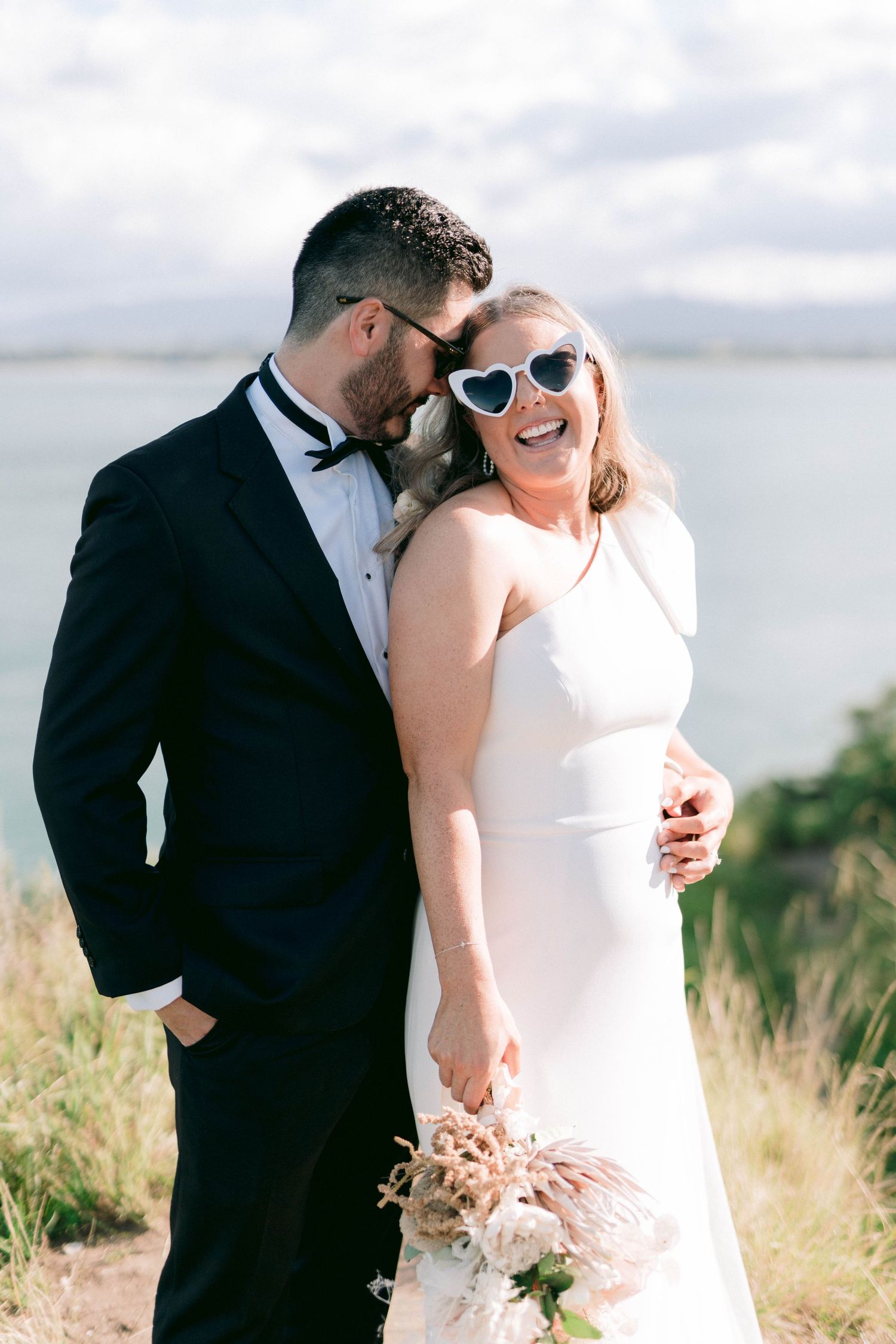 A joyful moment captured between a couple in wedding attire with sunglasses on a hilltop by the water.