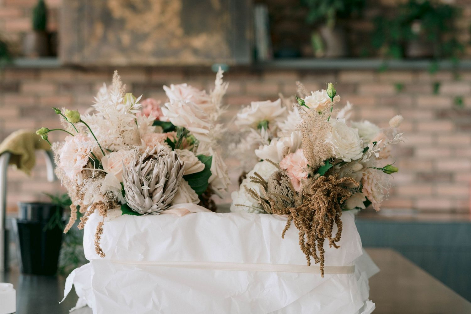 A rustic floral arrangement with soft pink and white flowers in a white container sits against a blurred background.