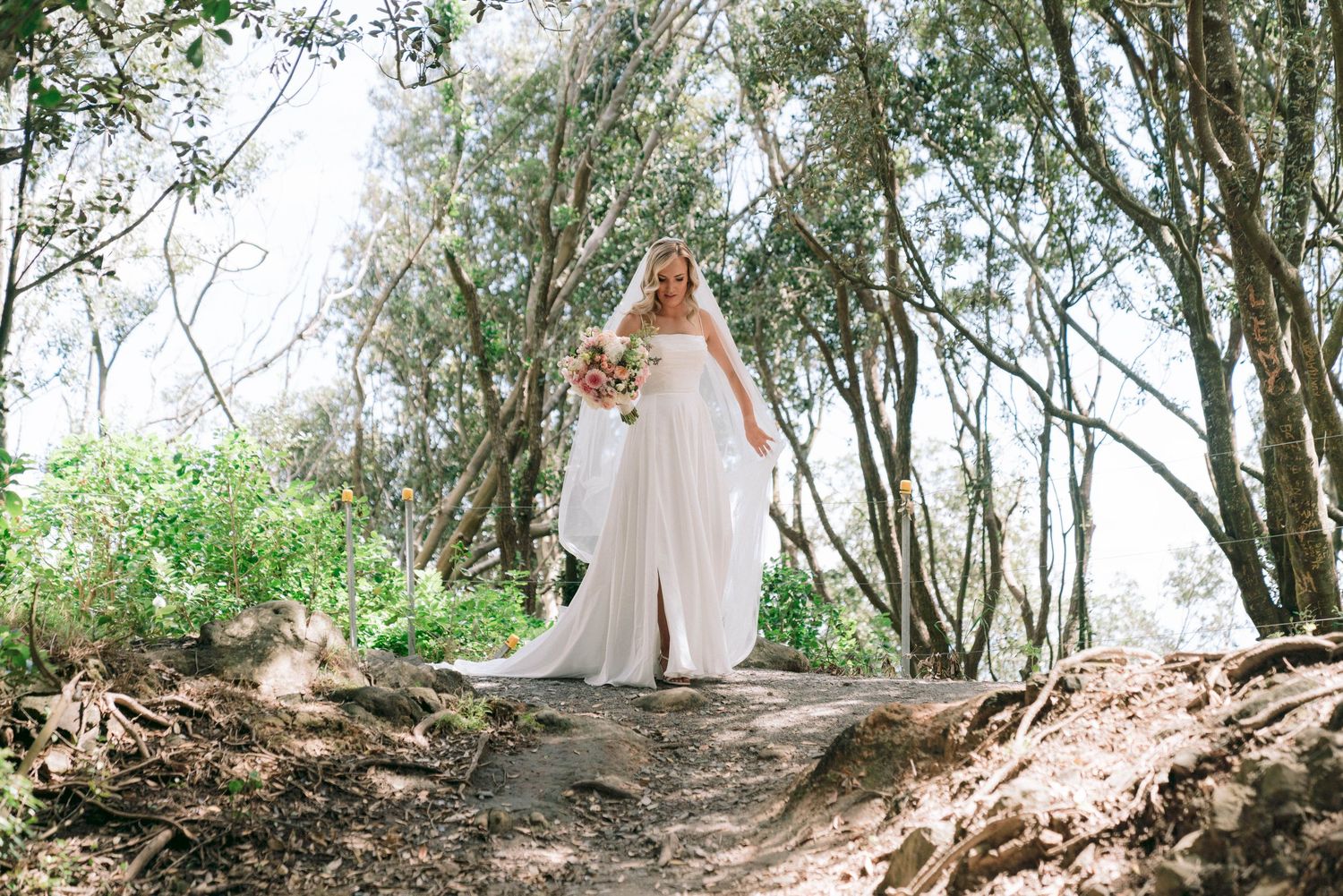 A bride in a white wedding gown walks along a forest path surrounded by tall trees and dappled sunlight.