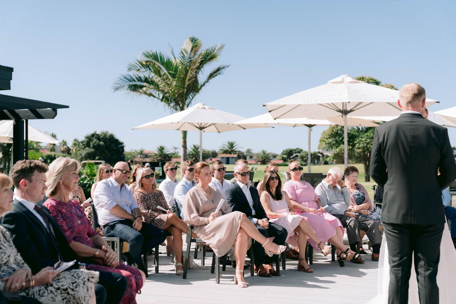 Wedding guests seated under white umbrellas at an elegant outdoor reception with palm trees.