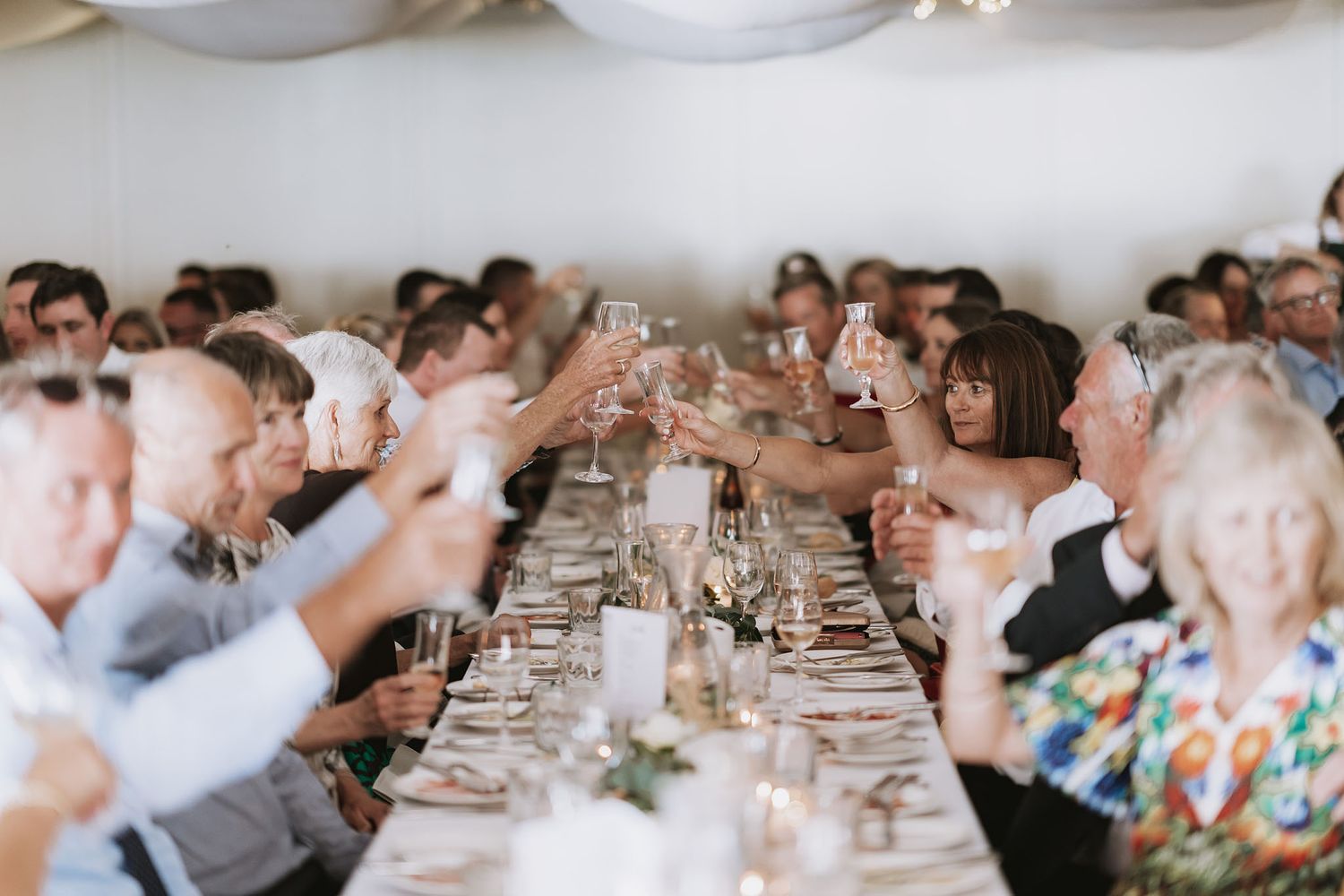 Wedding guests raise their glasses in a toast at a long banquet table under a white tent during a reception celebration.