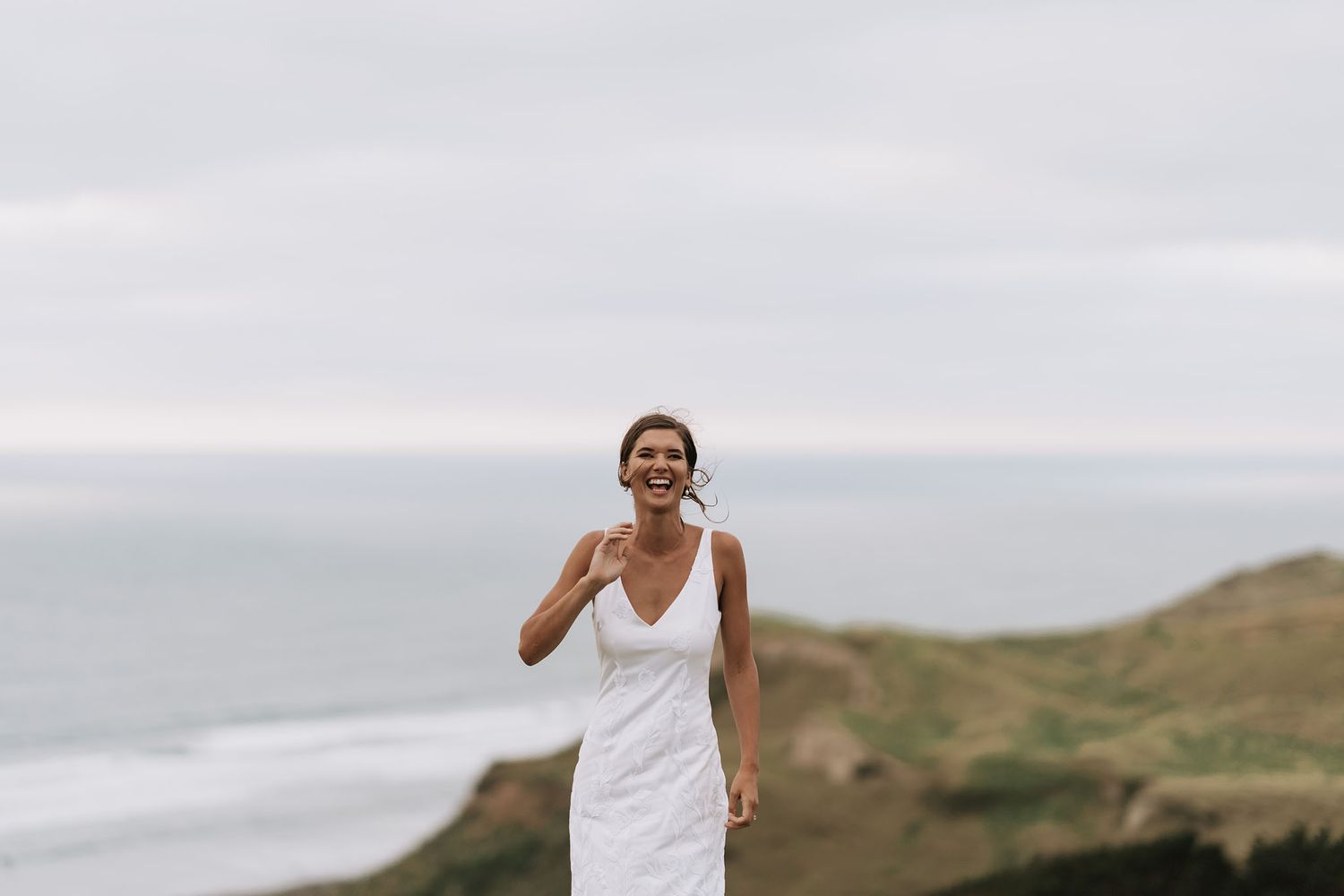 A person in a white dress runs joyfully along a scenic coastal cliff path overlooking the ocean on an overcast day.
