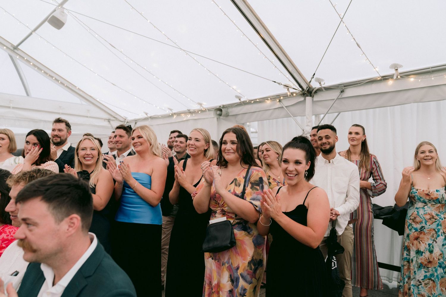 Wedding guests cheer and dance during a festive reception under a white tent canopy.