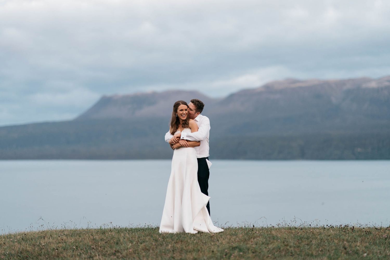 Two figures in white embrace lovingly on a scenic cliffside overlooking a serene mountain lake in the distance.