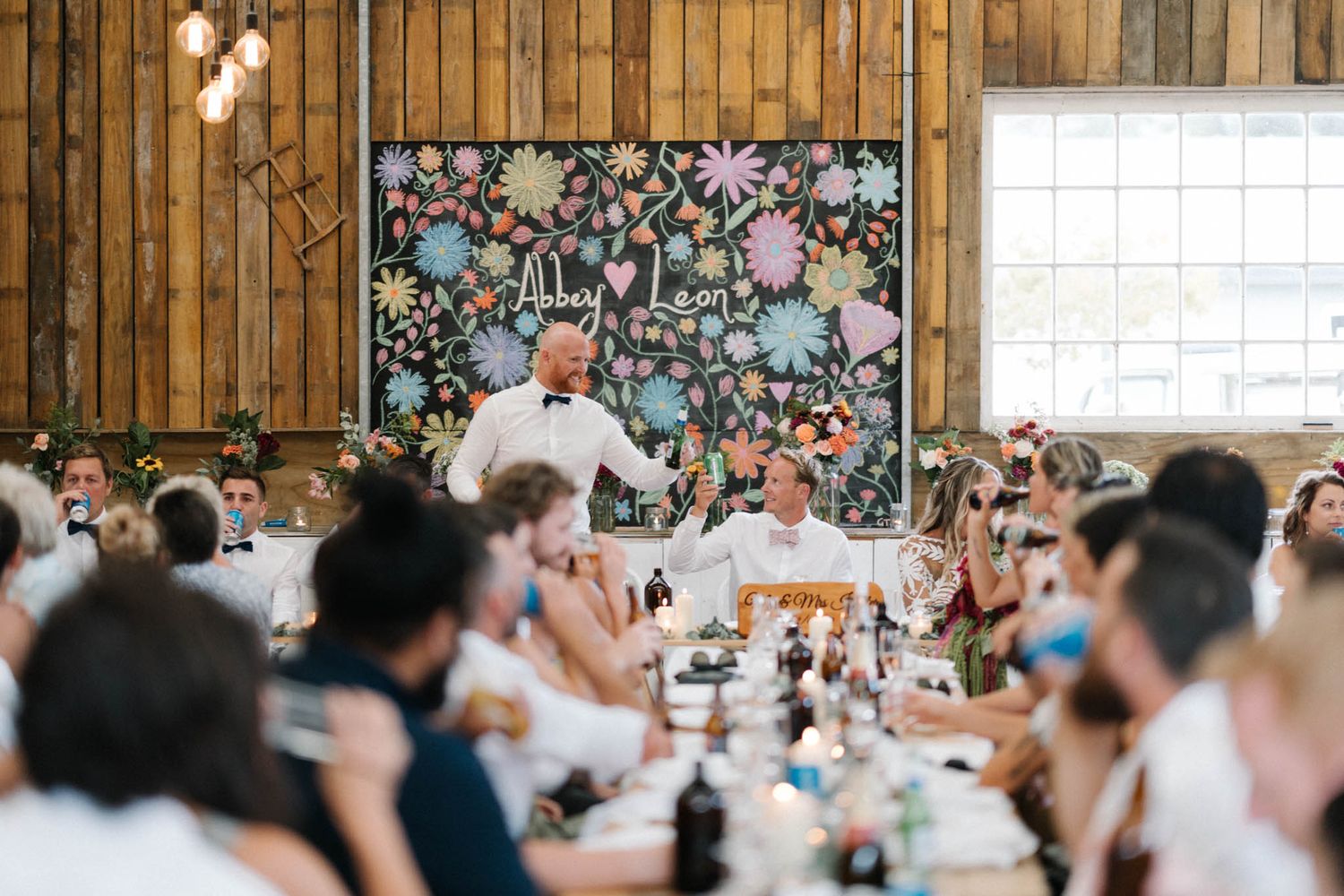 Guests mingle and celebrate at a rustic barn wedding reception with twinkling lights and wooden beams overhead.