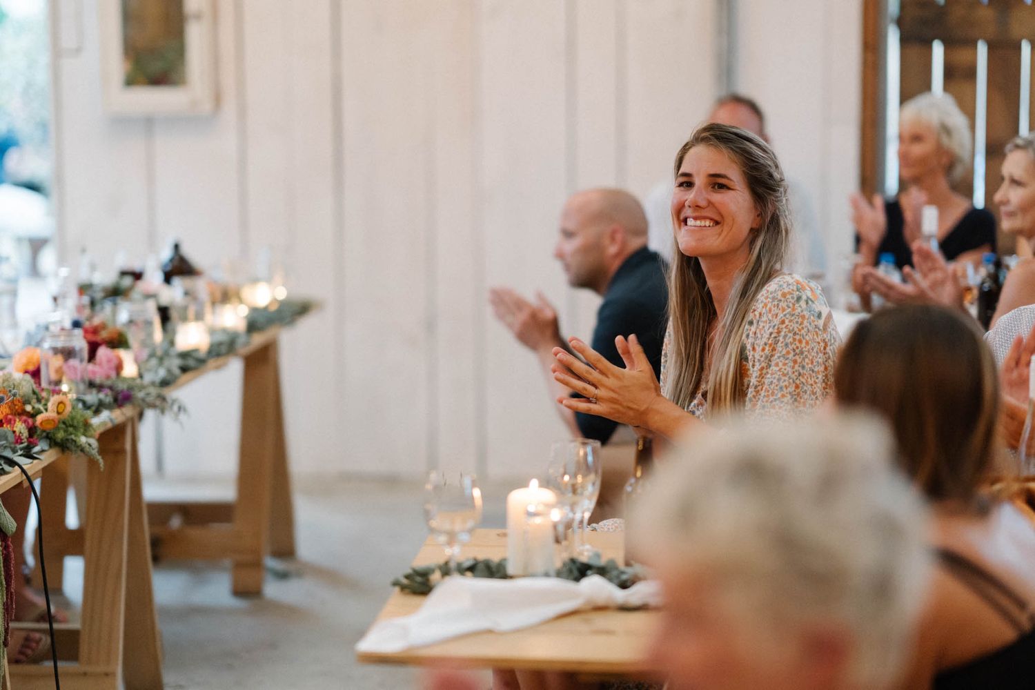 Wedding guests seated at long tables during an elegant indoor reception with candlelight and white decor.
