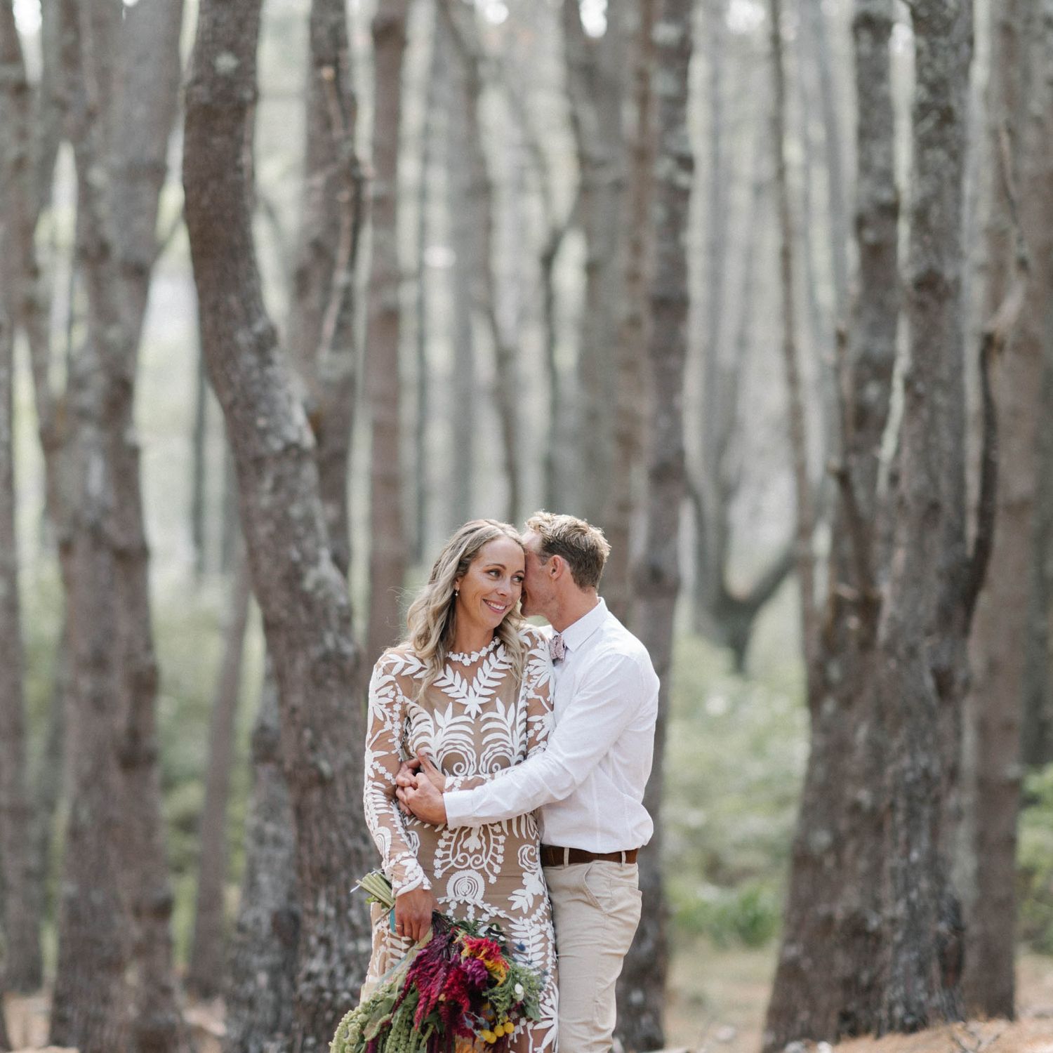 A couple embraces intimately in a forest of tall trees while holding a vibrant red bouquet.