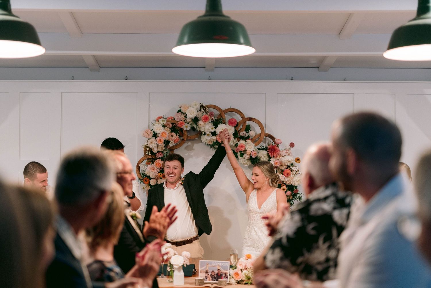 Wedding celebration under a floral arch with guests cheering during an indoor reception.
