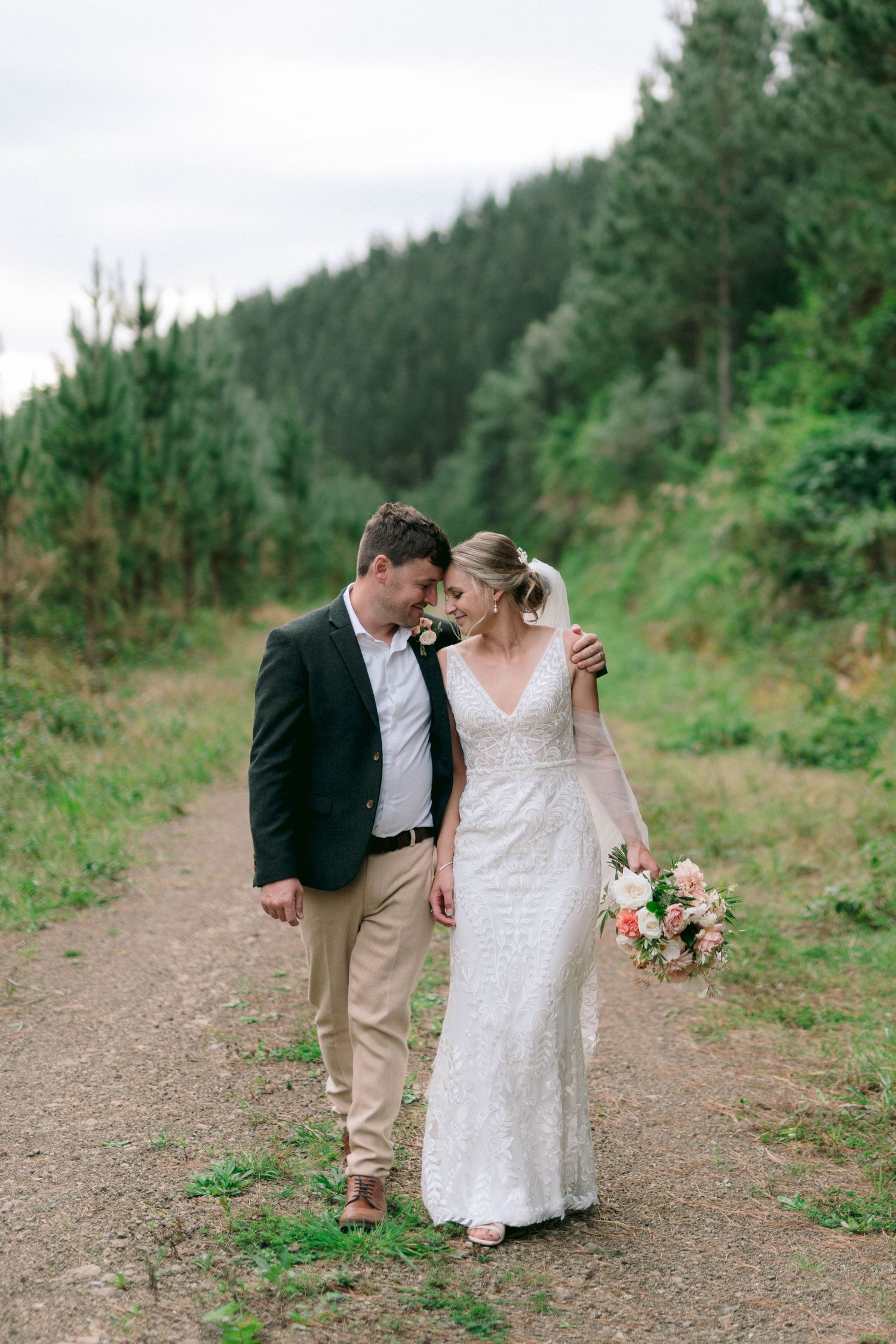 A romantic outdoor wedding photoshoot shows a couple walking on a dirt path surrounded by greenery.