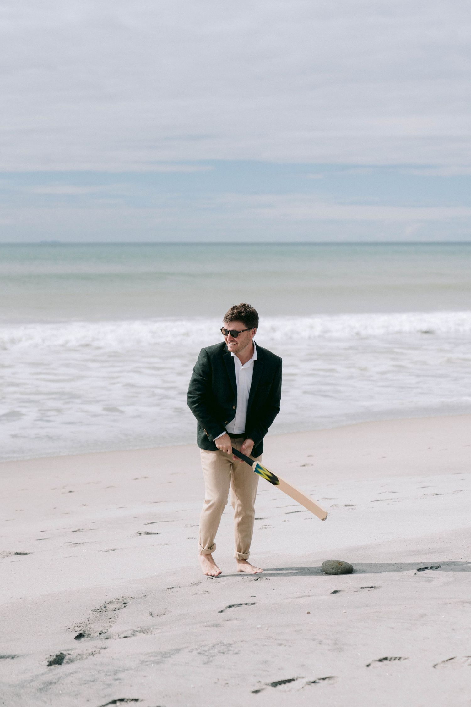 A person in a navy jacket and khaki pants walks on a serene beach holding a wooden baseball bat.