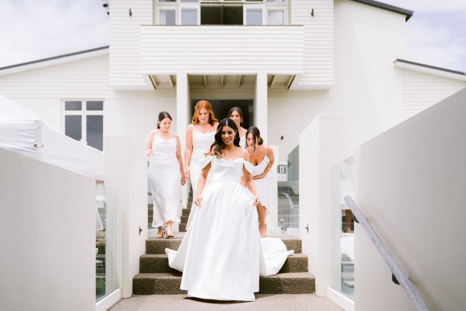 An elegant white wedding party descends the stairs of a modern white coastal building on a bright sunny day.