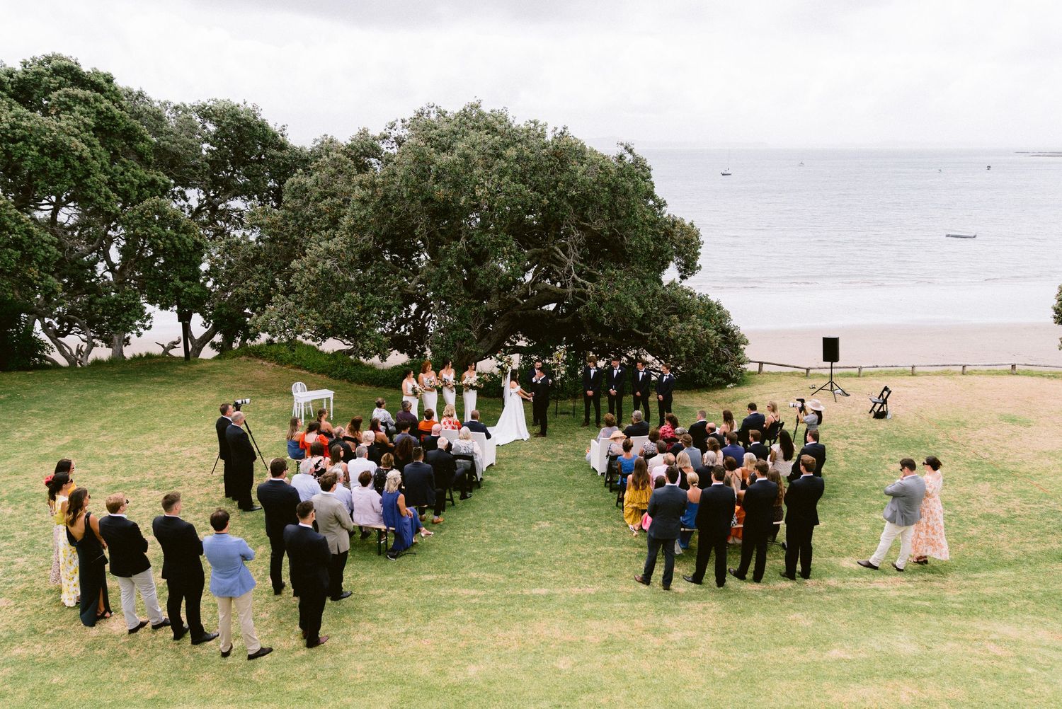 Outdoor wedding ceremony setup with guests seated on lawn chairs under large trees by the ocean.