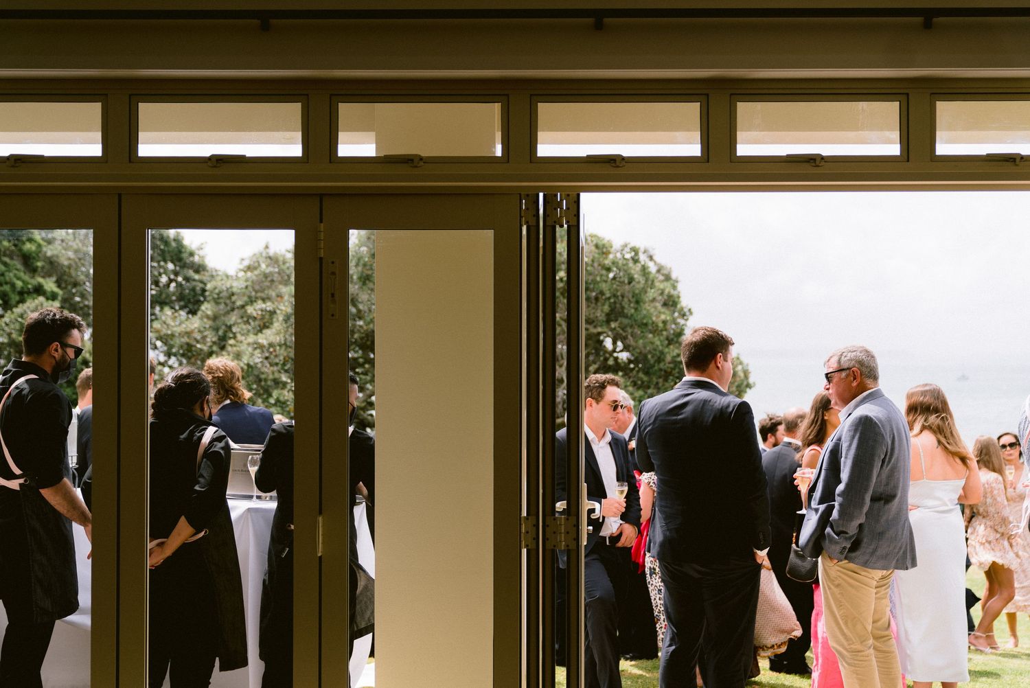 Wedding guests mingling in doorway between indoor and outdoor spaces at reception venue.