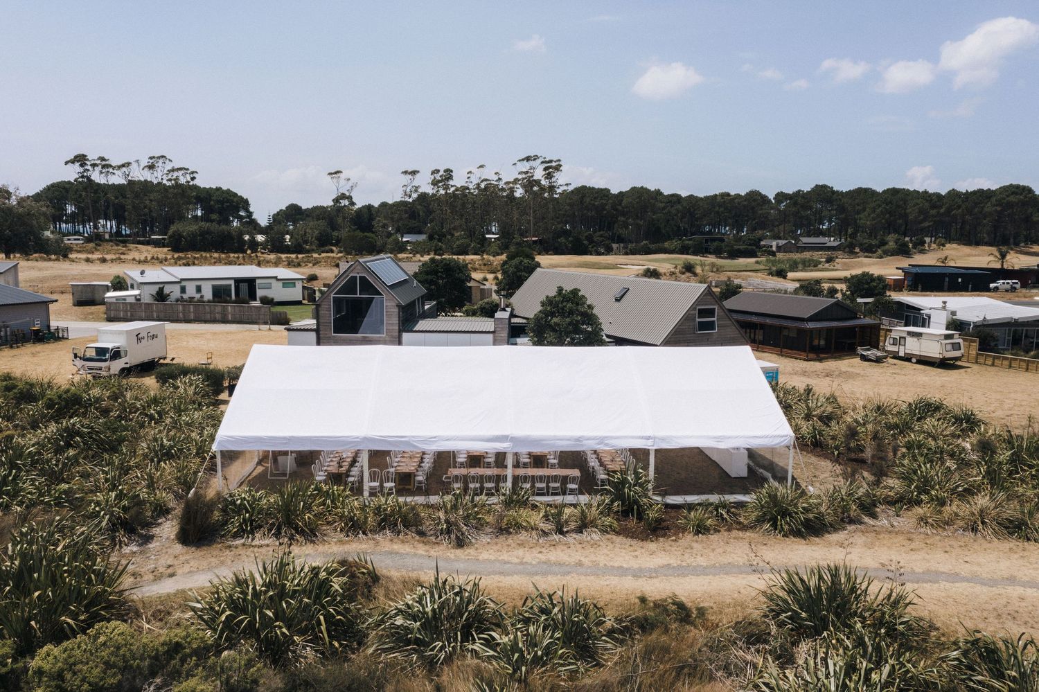 Large white marquee tent set up in a rural setting with scattered buildings and eucalyptus trees in the background.