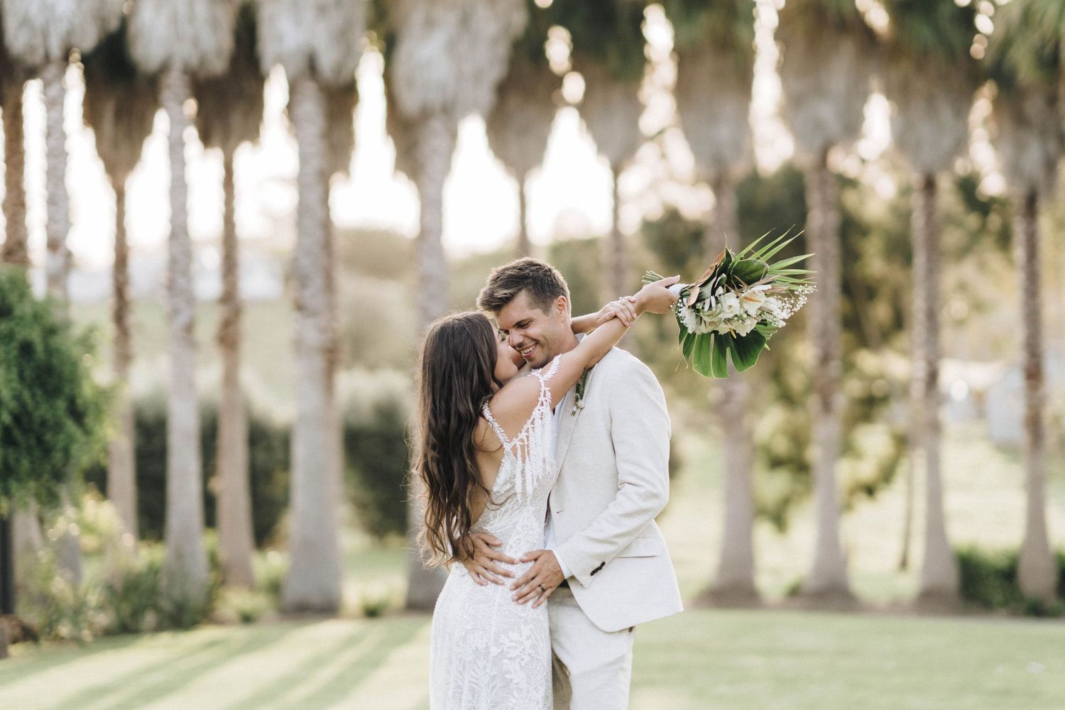 Romantic outdoor wedding photography session between palm trees with natural sunlight filtering through.