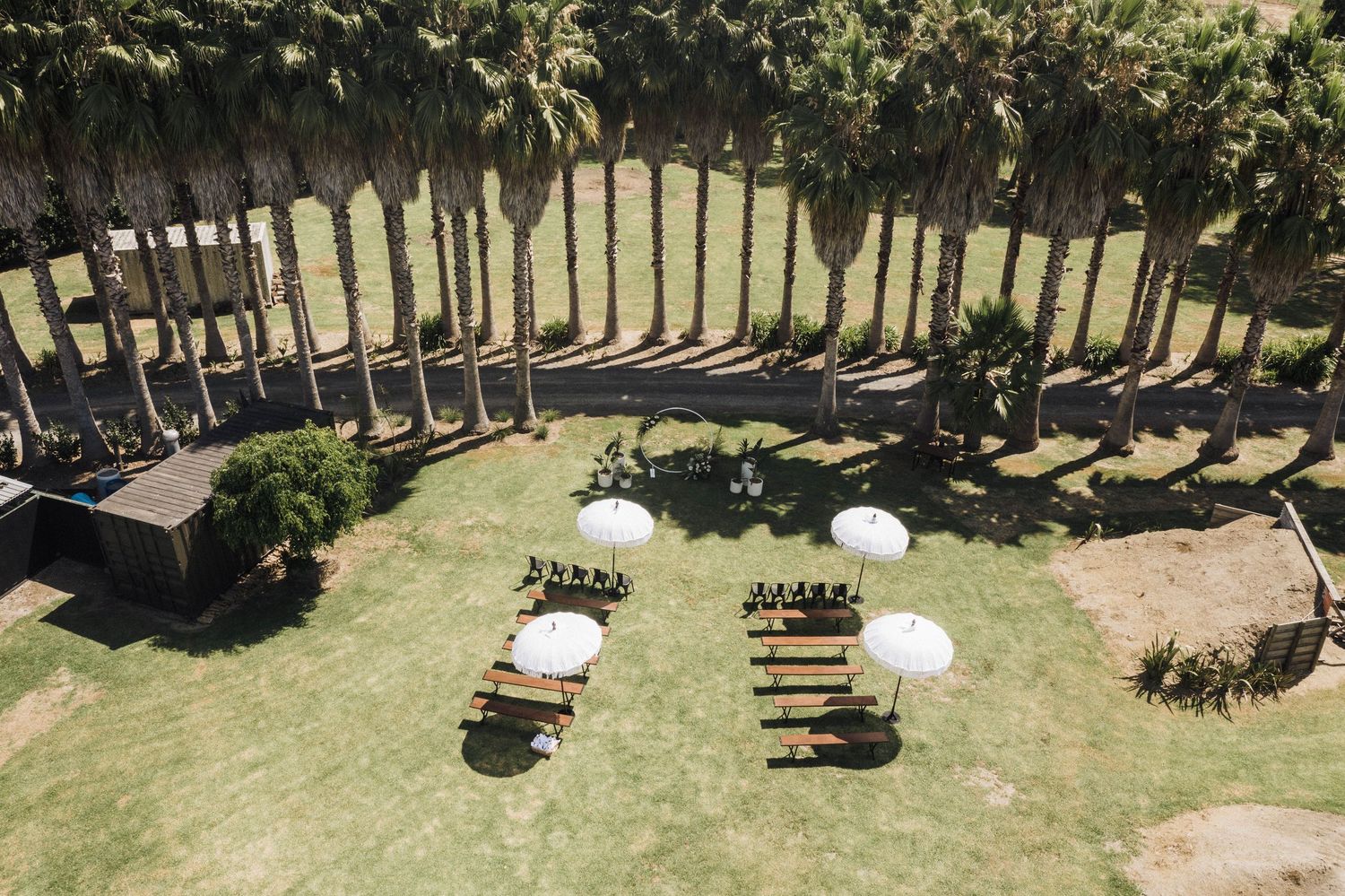 Aerial view of an outdoor wedding setup with wooden benches and white umbrellas arranged on a circular lawn surrounded by trees.