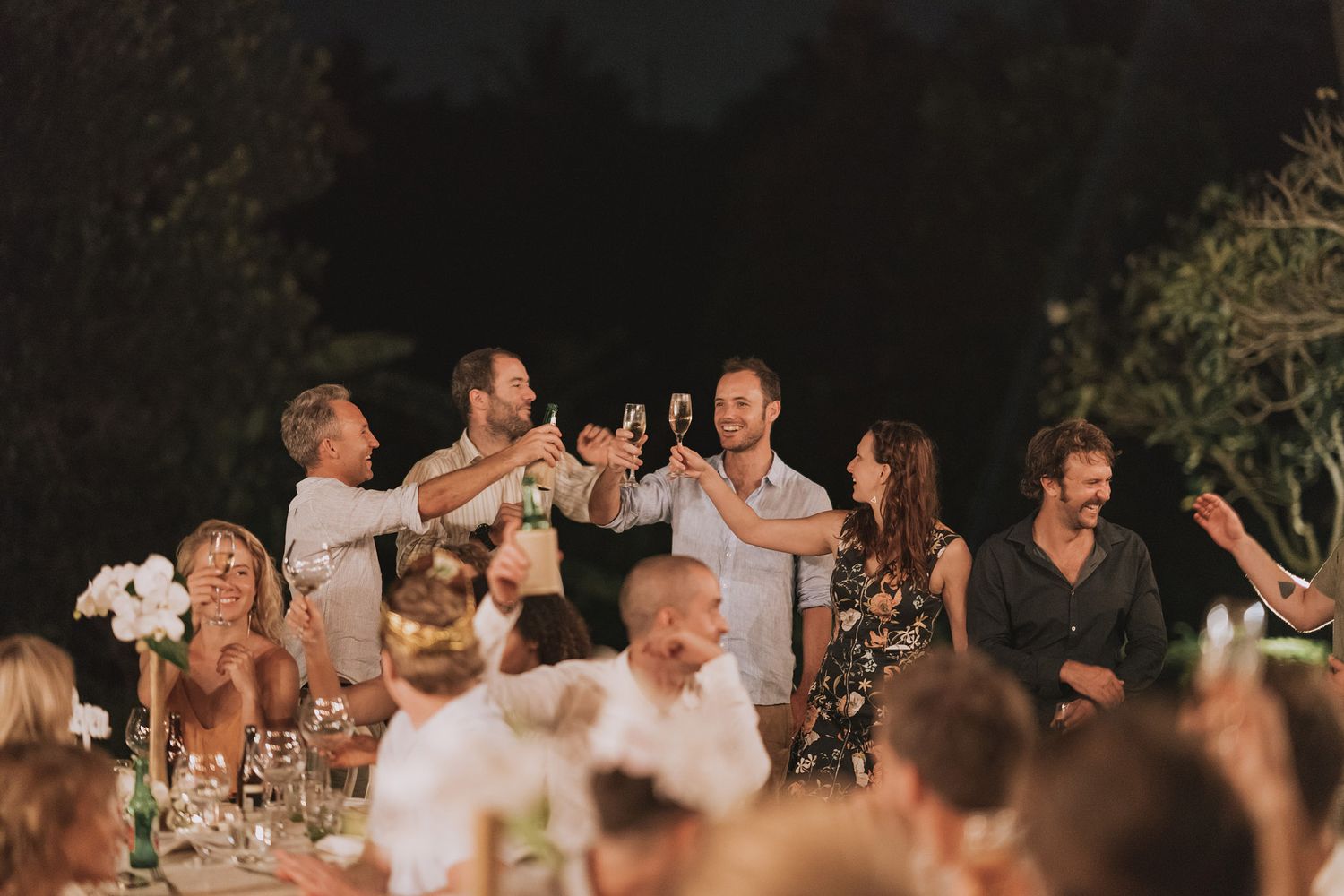 Wedding guests raising glasses in a toast during outdoor evening reception.