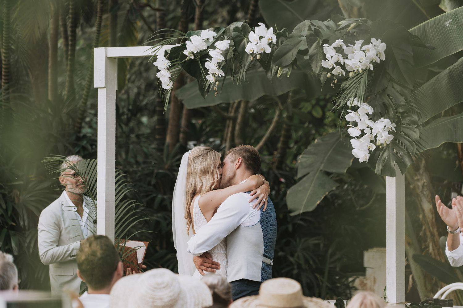 Romantic wedding ceremony setup with white archway framed by tropical greenery and flowers.