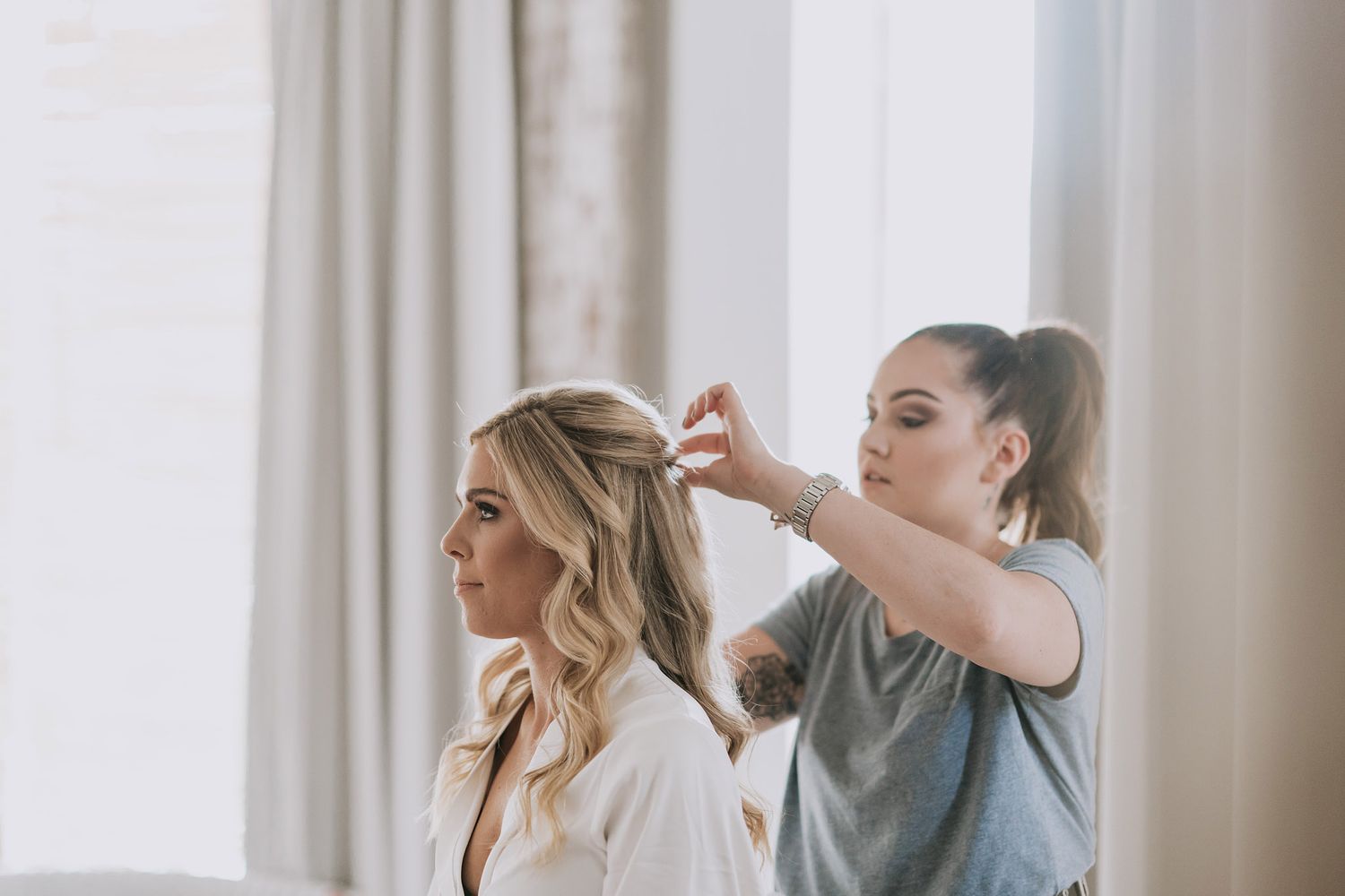 A hairstylist works on styling a client's blonde hair in a bright, minimalist room with white curtains.