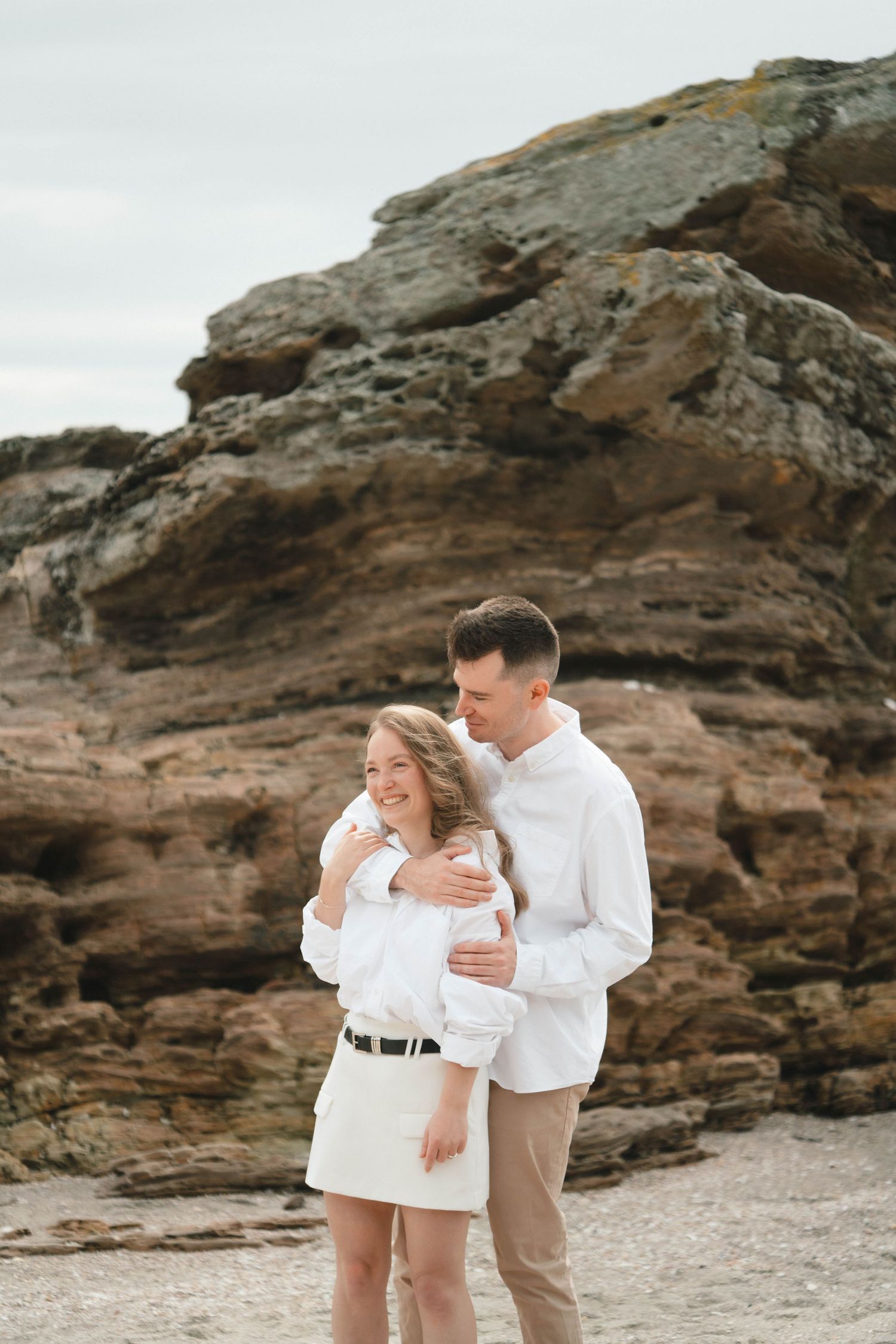A couple in white clothing embraces against a backdrop of rugged coastal cliffs.
