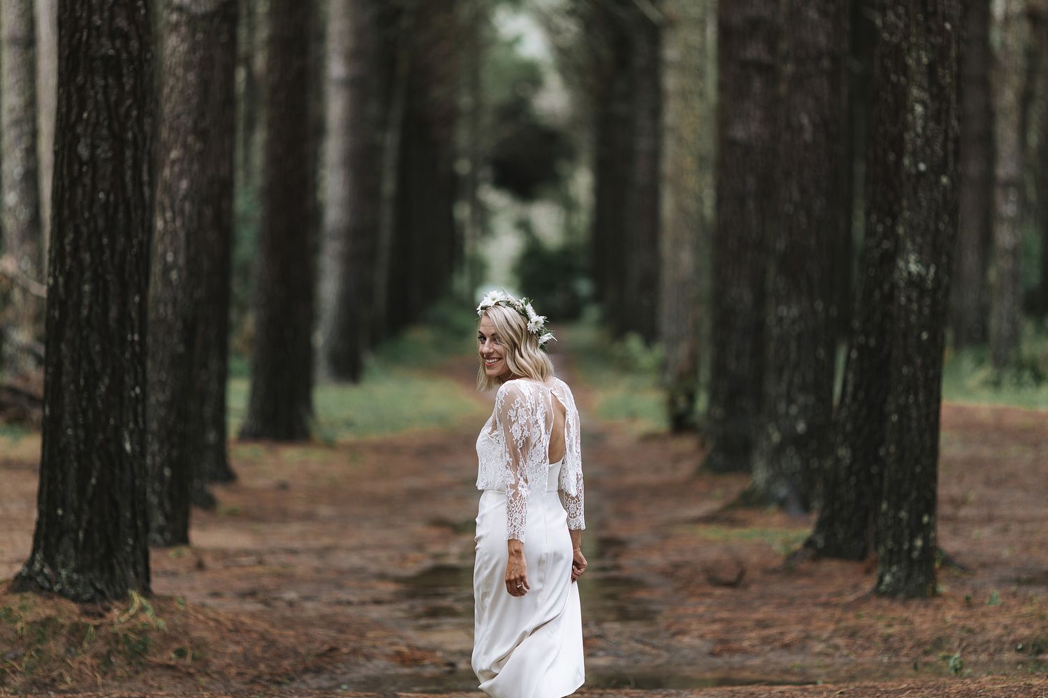 A person in a white dress stands in a serene pine forest path, creating an ethereal woodland scene.