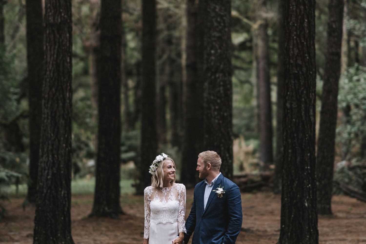 A couple dressed in wedding attire standing together in a moody evergreen forest.