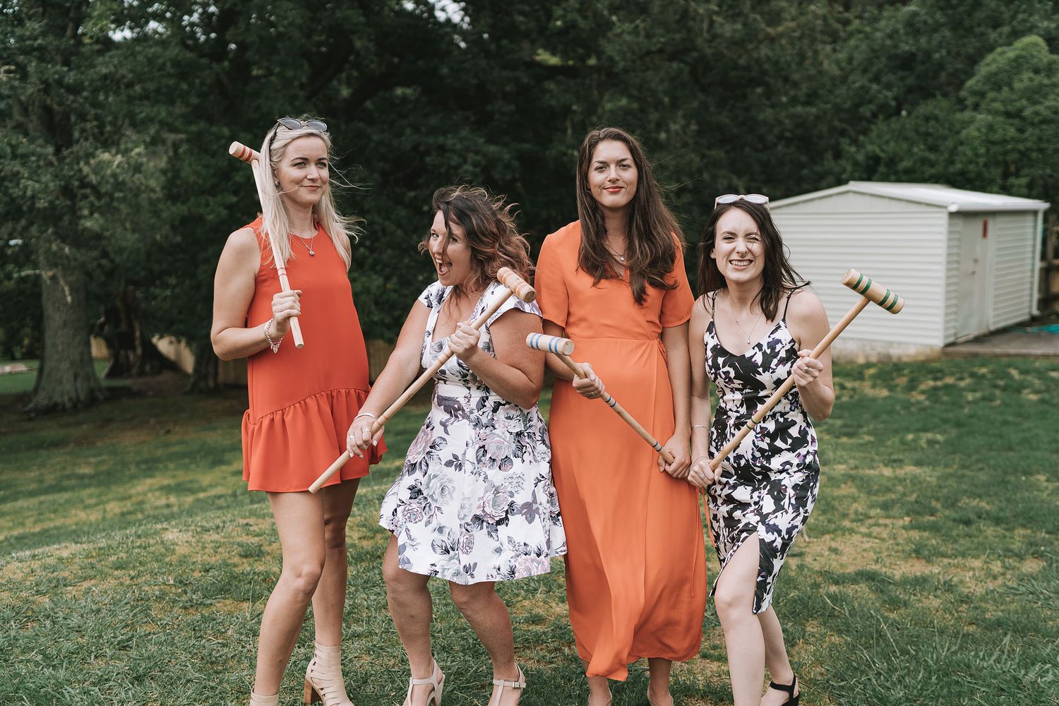 Four people in summer dresses stand together on a lawn holding gardening tools and smiling.