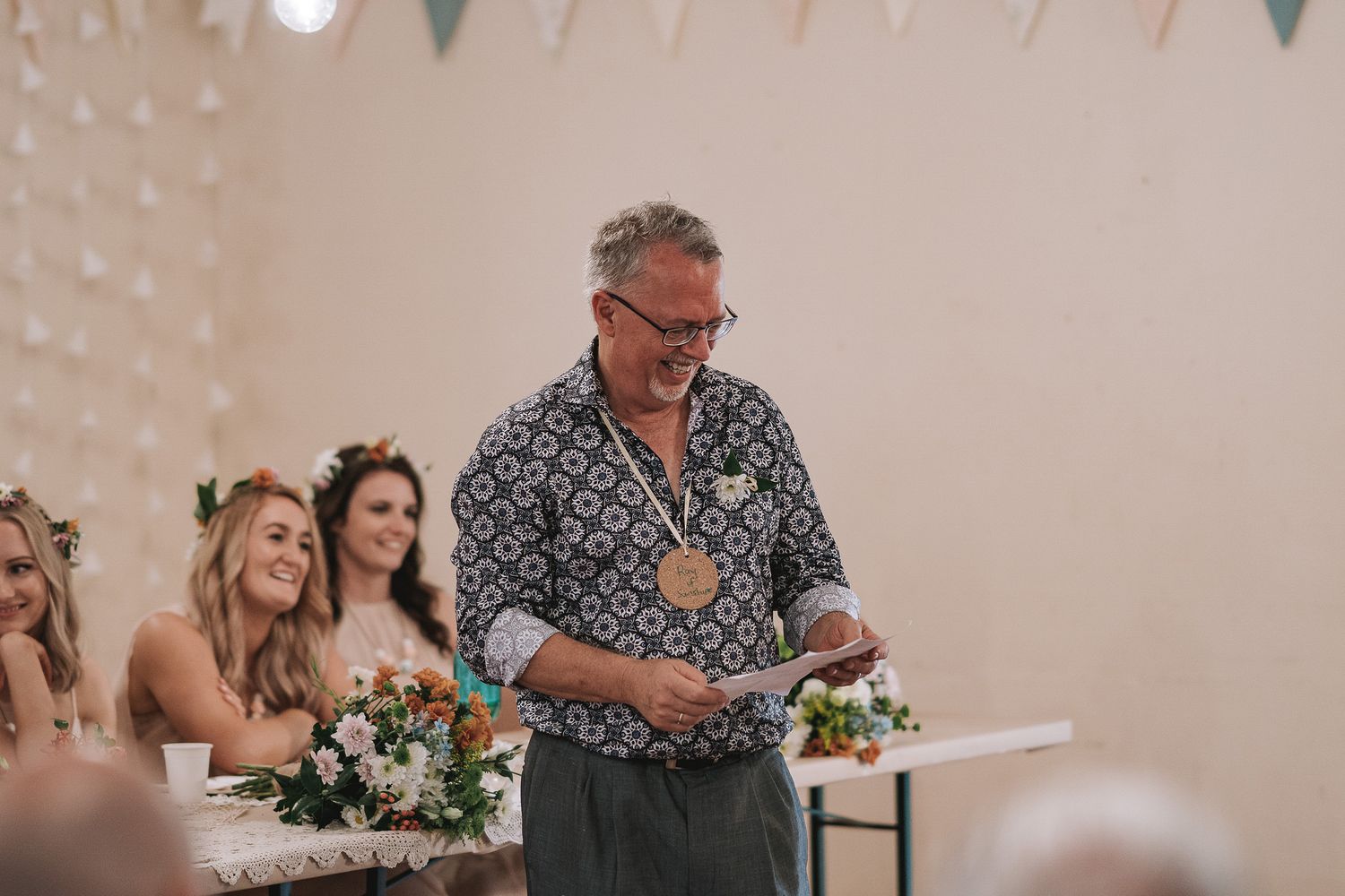 A person in a patterned button-up shirt gives a speech at a wedding reception with bridesmaids seated nearby.