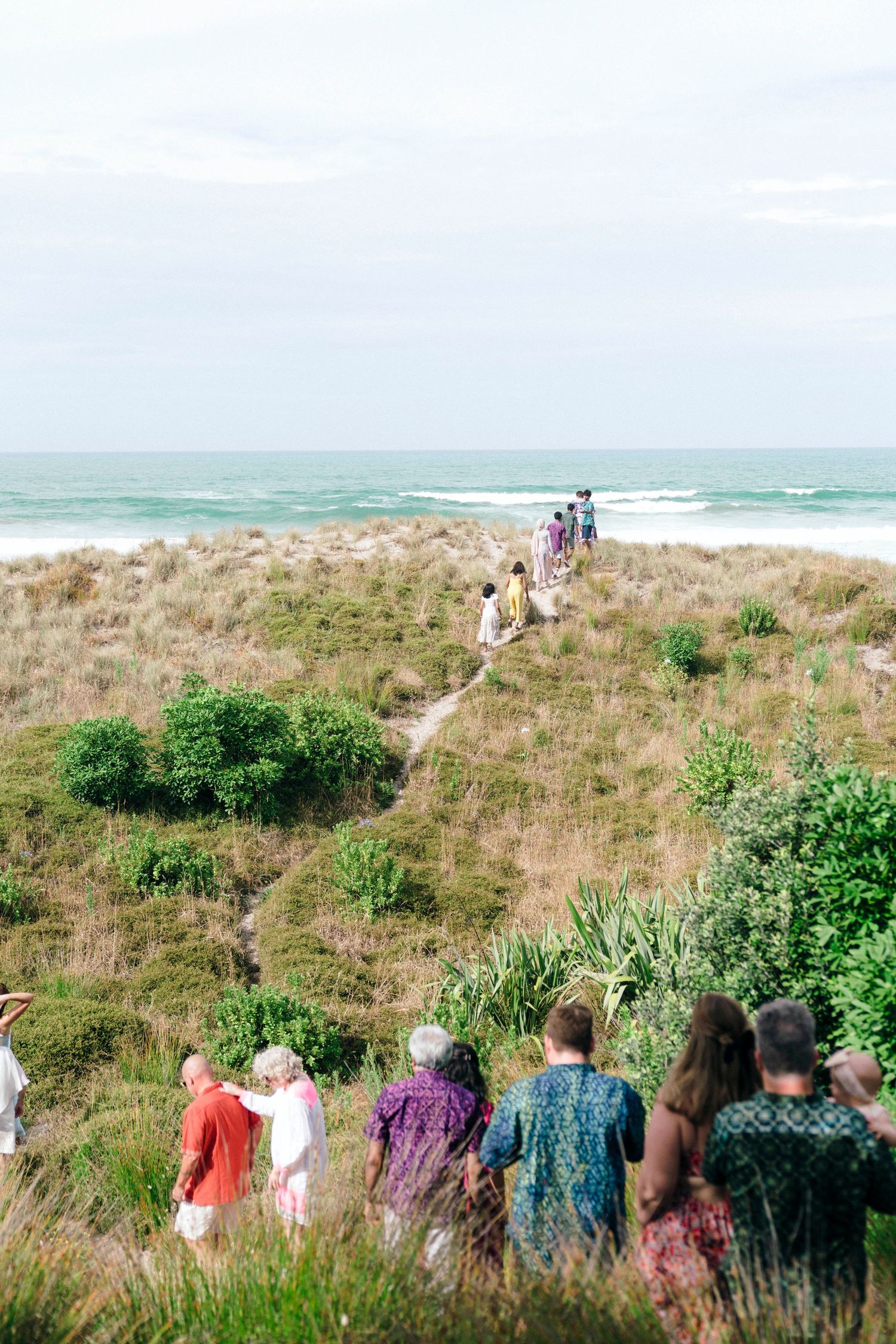 A group of wedding guests walking down a grassy coastal path toward the ocean on a bright day.
