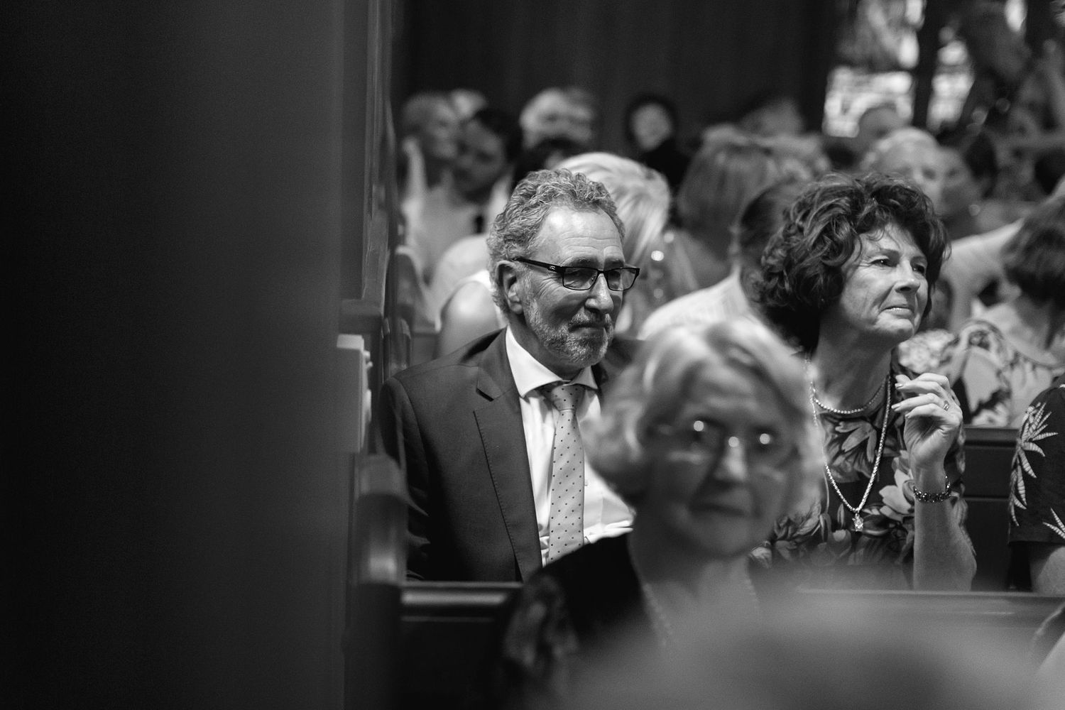 Black and white photograph showing people seated in an audience at an event, with some attendees smiling and engaged.