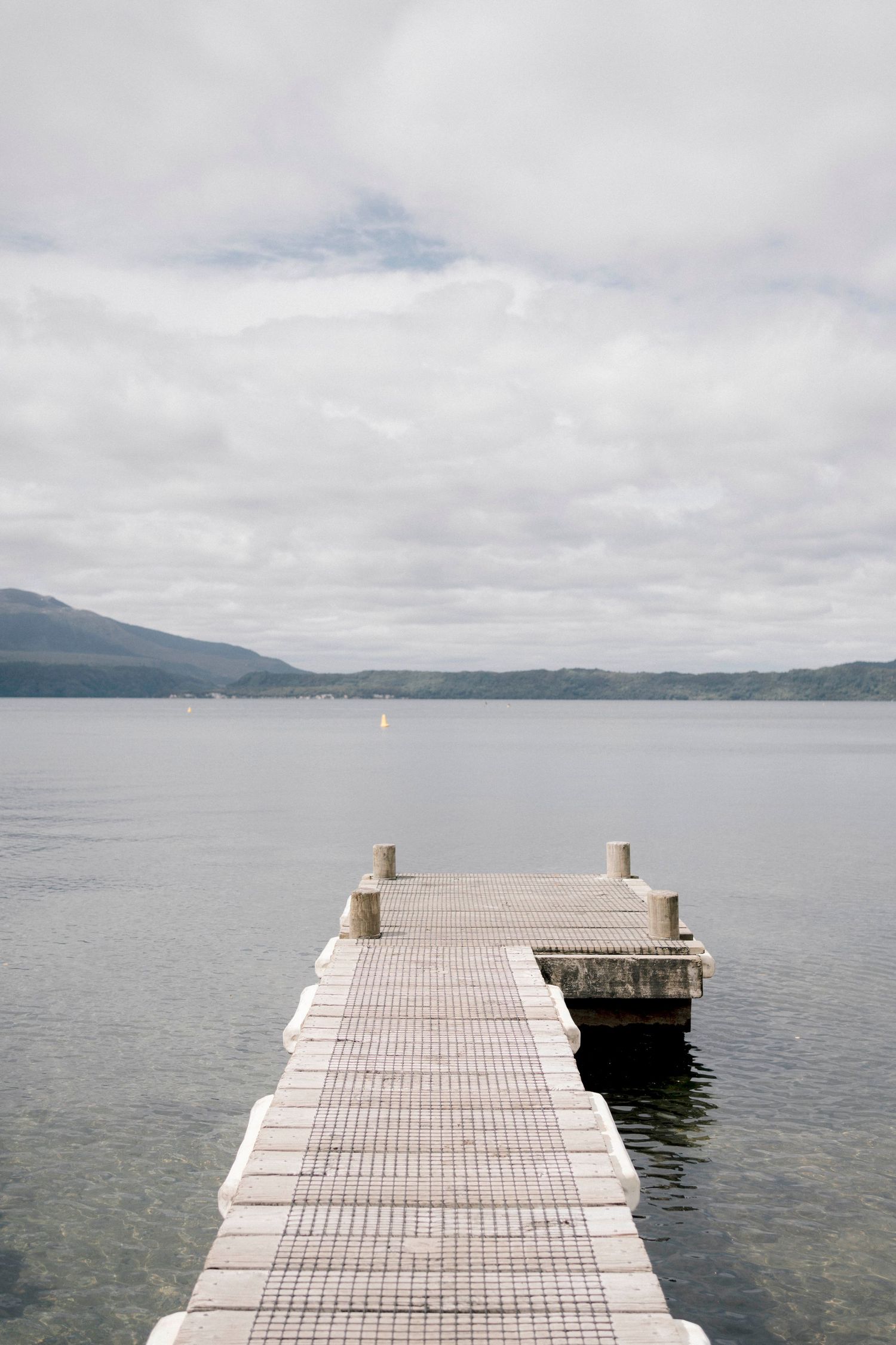 A wooden dock extends over calm lake waters with mountains and cloudy skies in the background.
