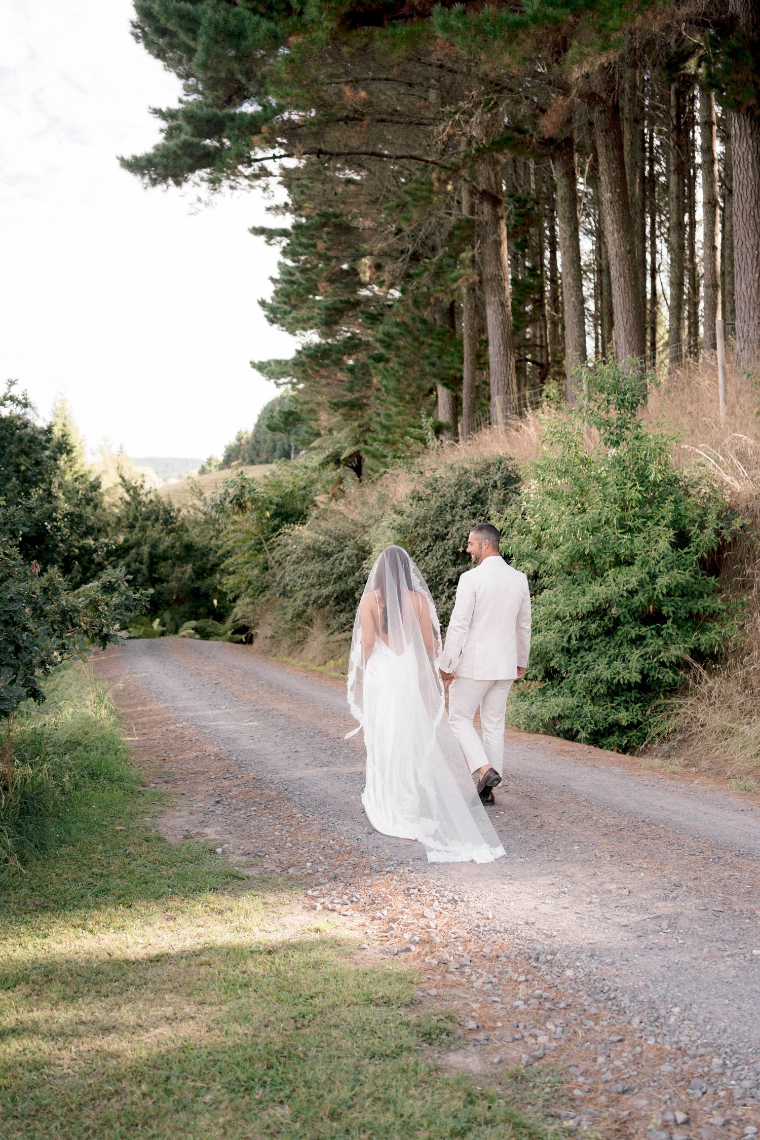 Couple walking down a forest path with bride in flowing white dress and veil.