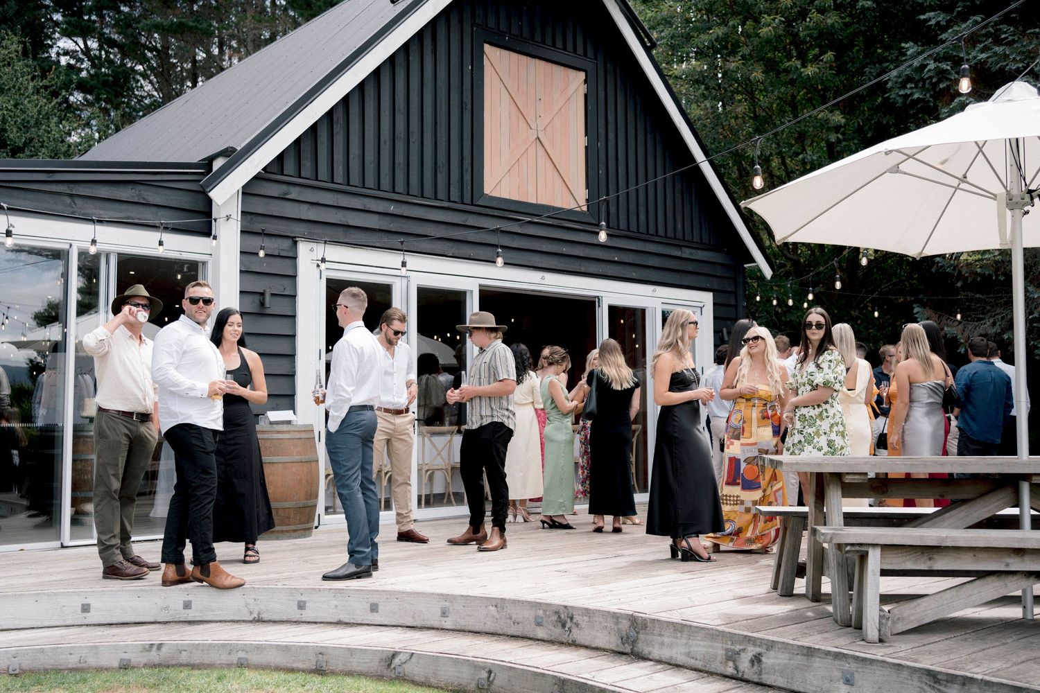 Wedding guests mingle outside a modern black barn venue with white umbrellas.