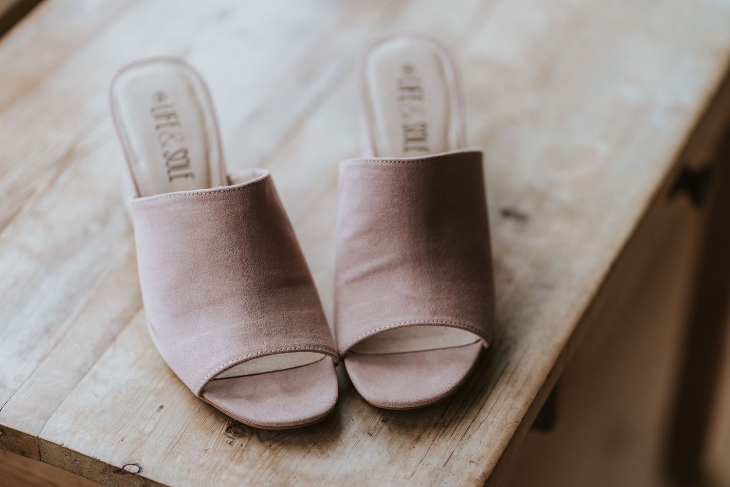 A pair of blush pink suede mule sandals with open toes sits on a light wooden surface.