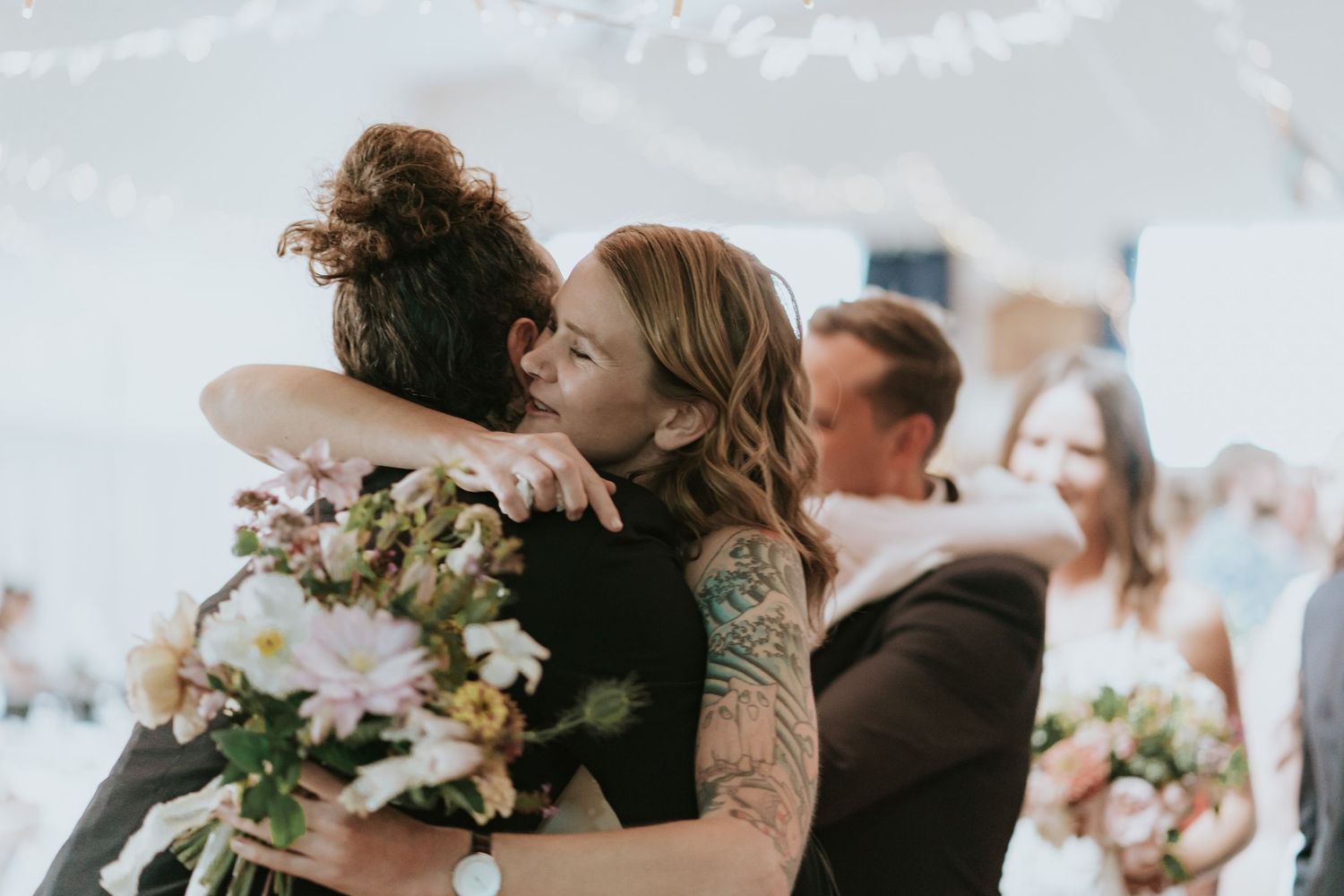 Two close friends sharing an emotional embrace at a wedding celebration while holding a floral bouquet.