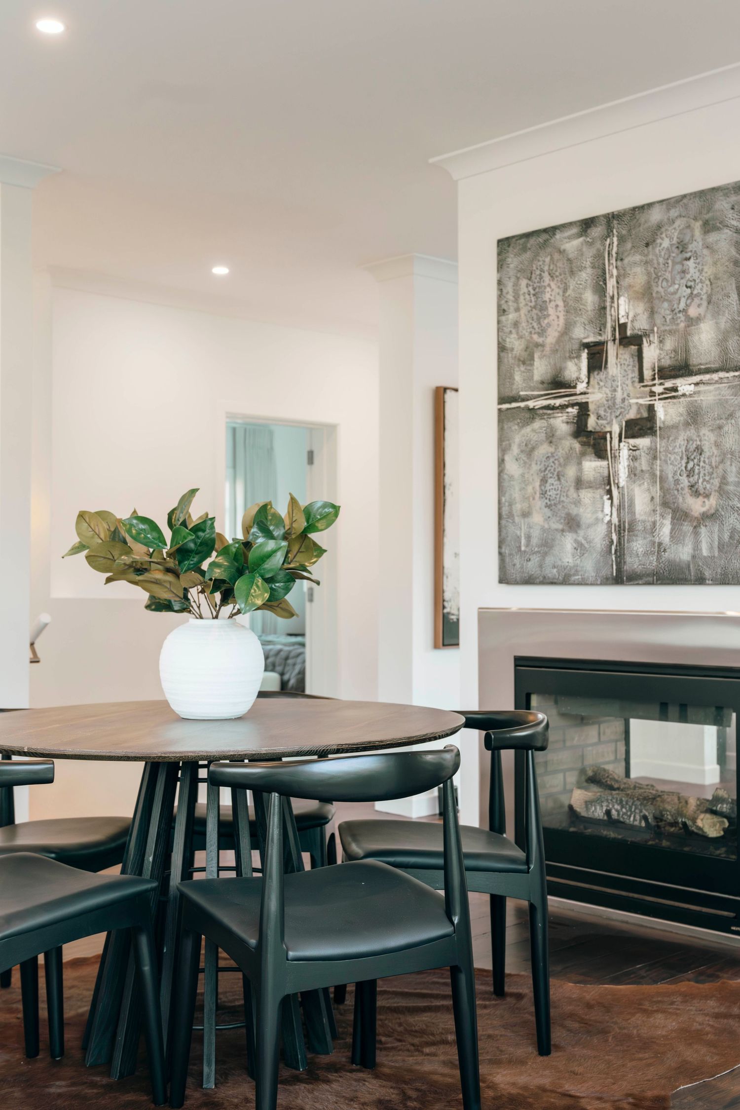 Modern dining area with round table, black chairs, white vase with greenery, and abstract cross artwork above fireplace.