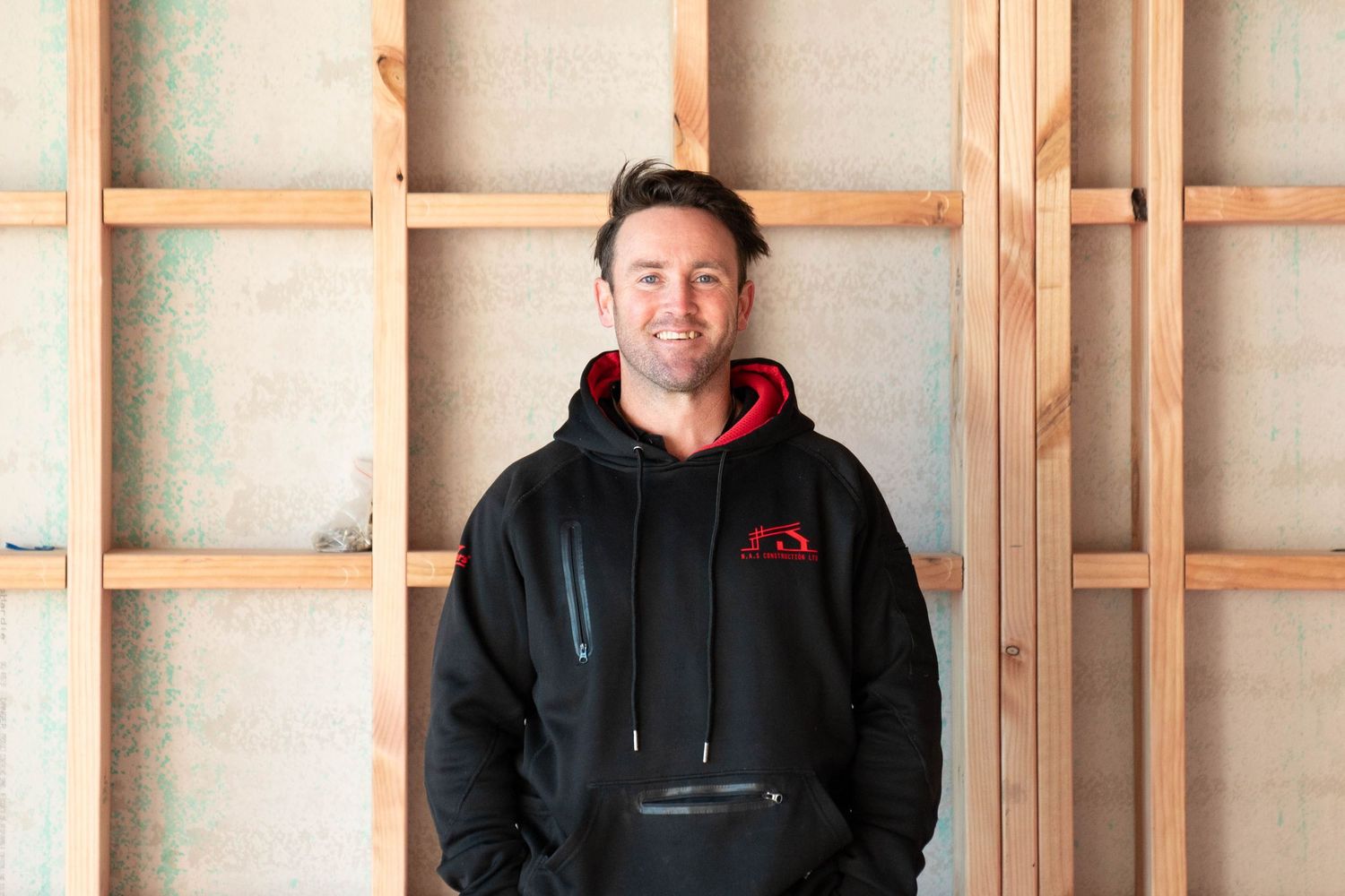 Person wearing a black hoodie with red logo standing against wooden framed wall with insulation material.