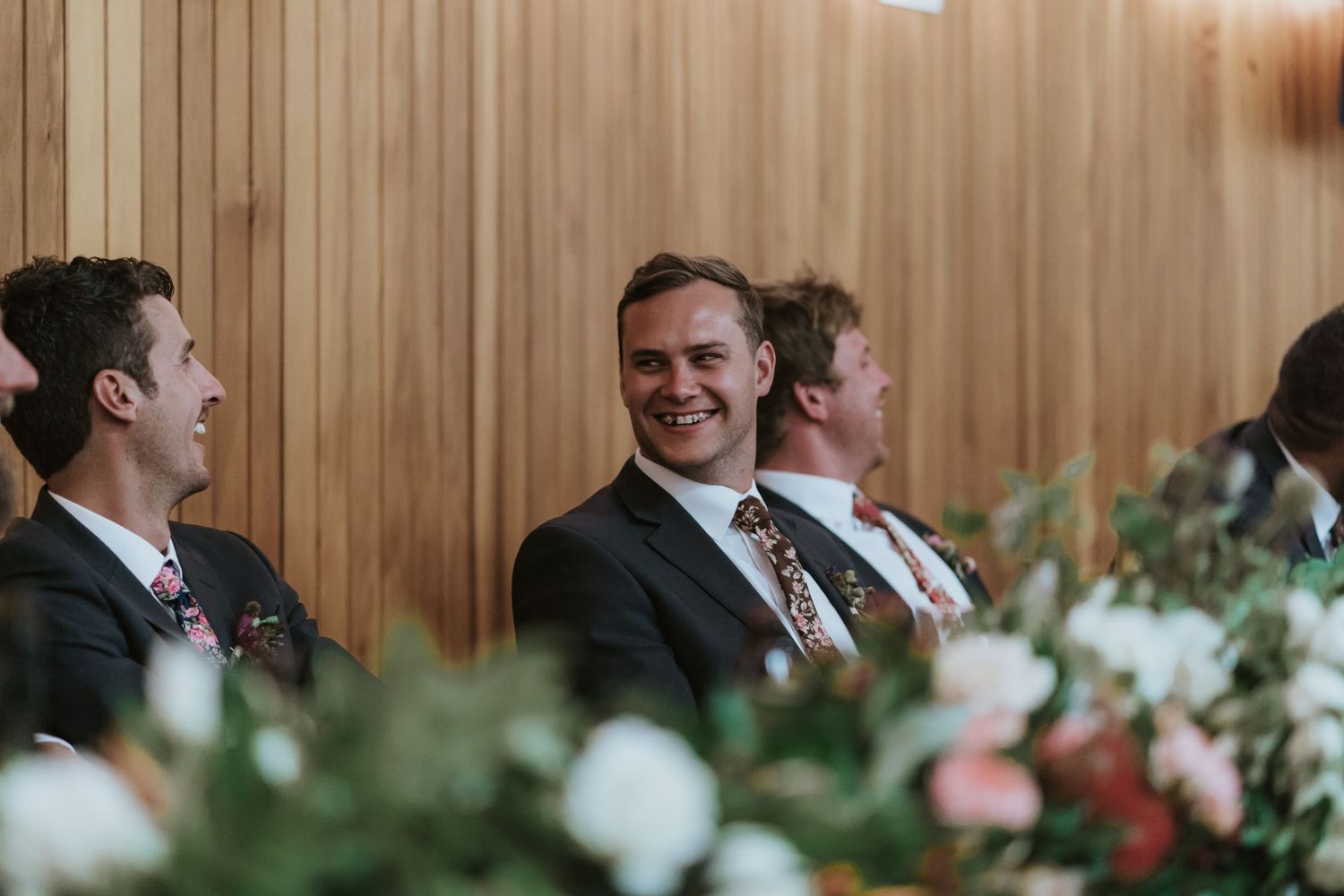 People seated at a formal event with decorative white and pink floral arrangements on wooden paneled walls.