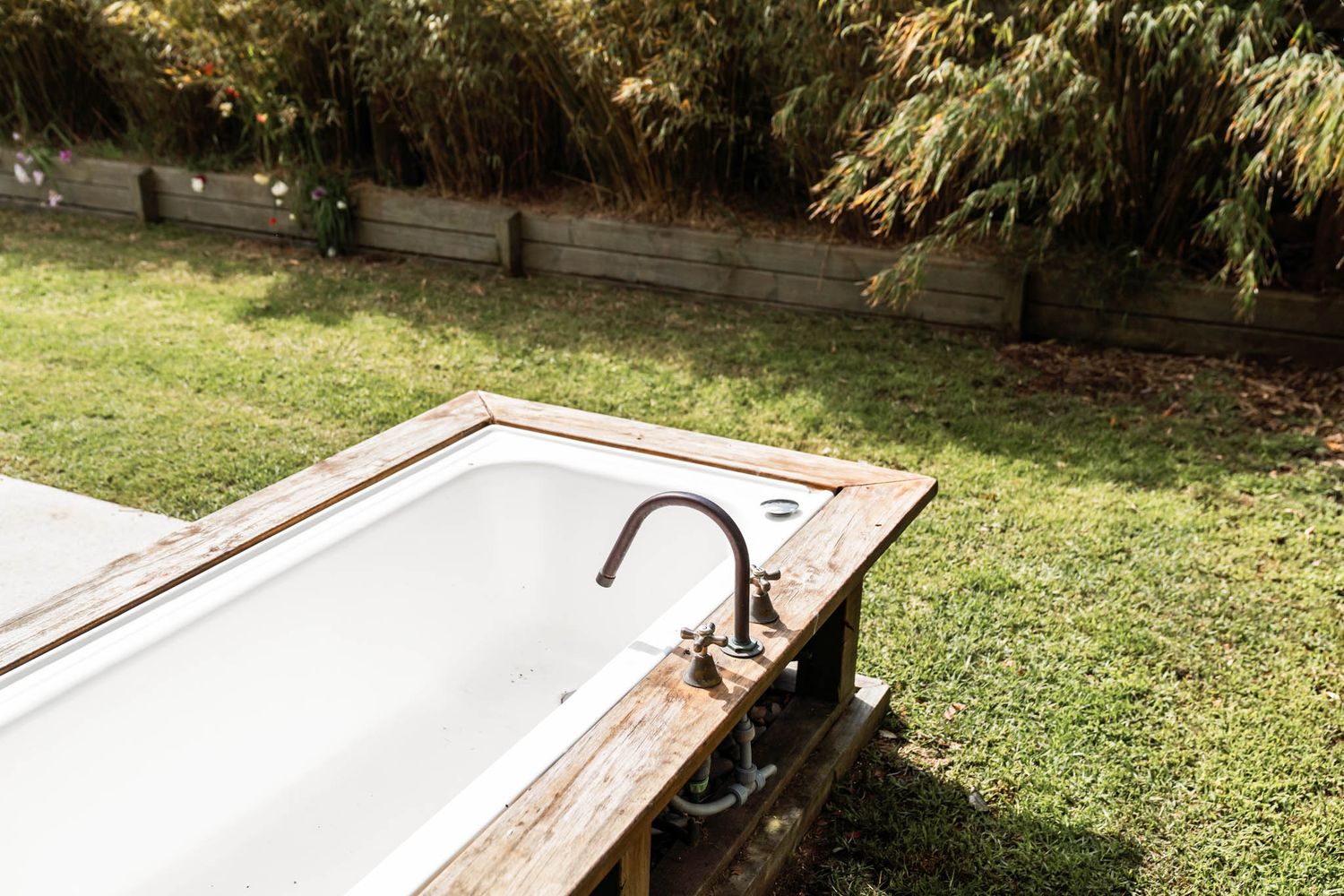 Rustic outdoor bathtub with faucet fixture placed on wooden platform in grassy garden setting.