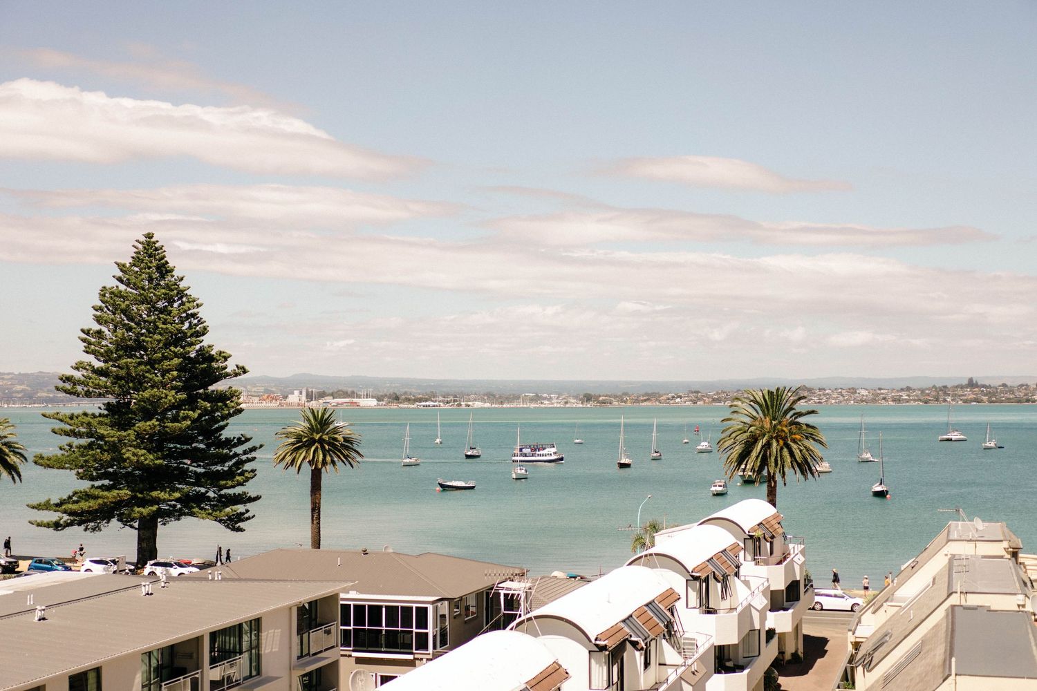 Boats dot the turquoise harbor waters along a coastal town with palm trees and white buildings under a cloudy sky.