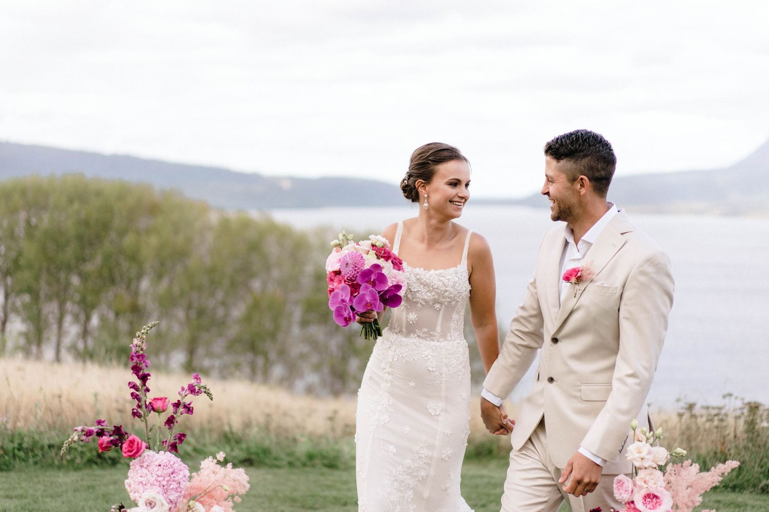 Couple embracing outdoors with pink floral arrangements against a scenic coastal backdrop.