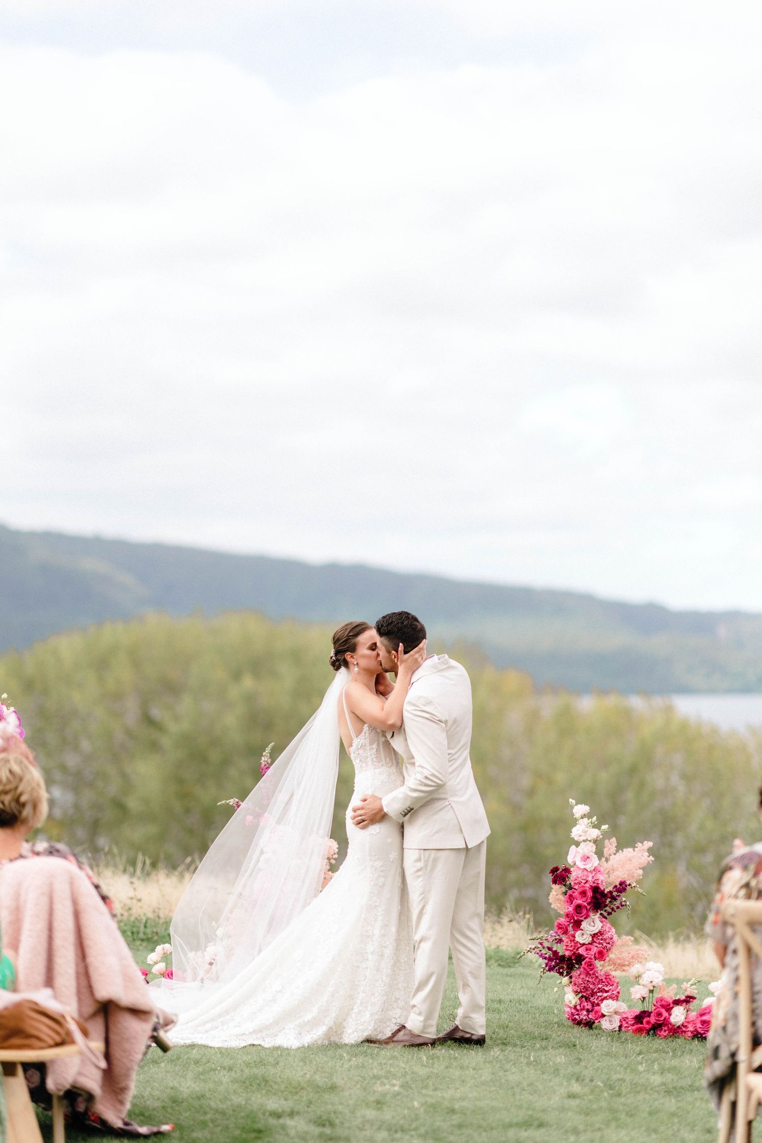 Outdoor wedding ceremony with pink floral arrangements near a lake and mountains.