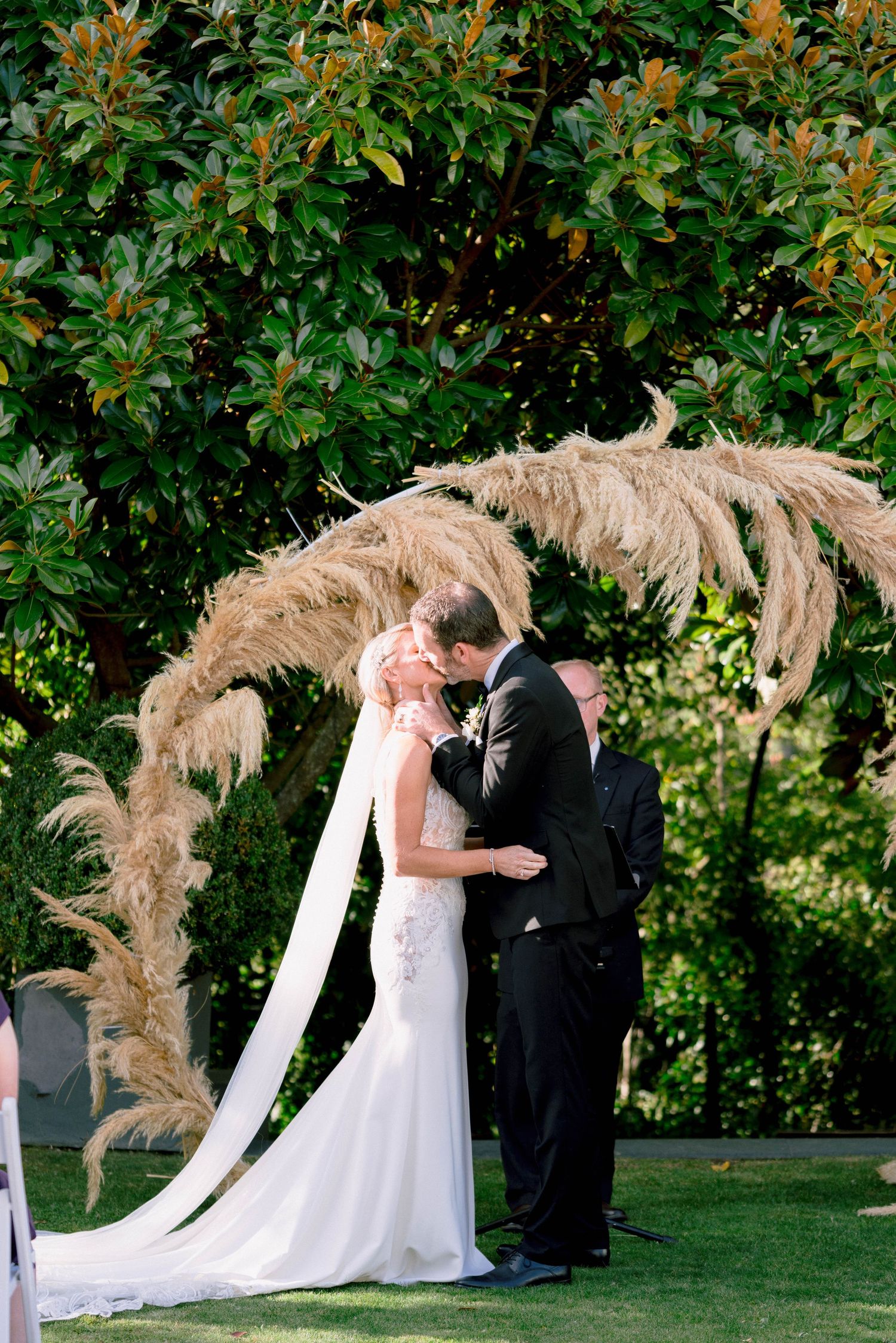 Multiple shots of a wedding ceremony under an outdoor arch decorated with pampas grass against lush greenery.