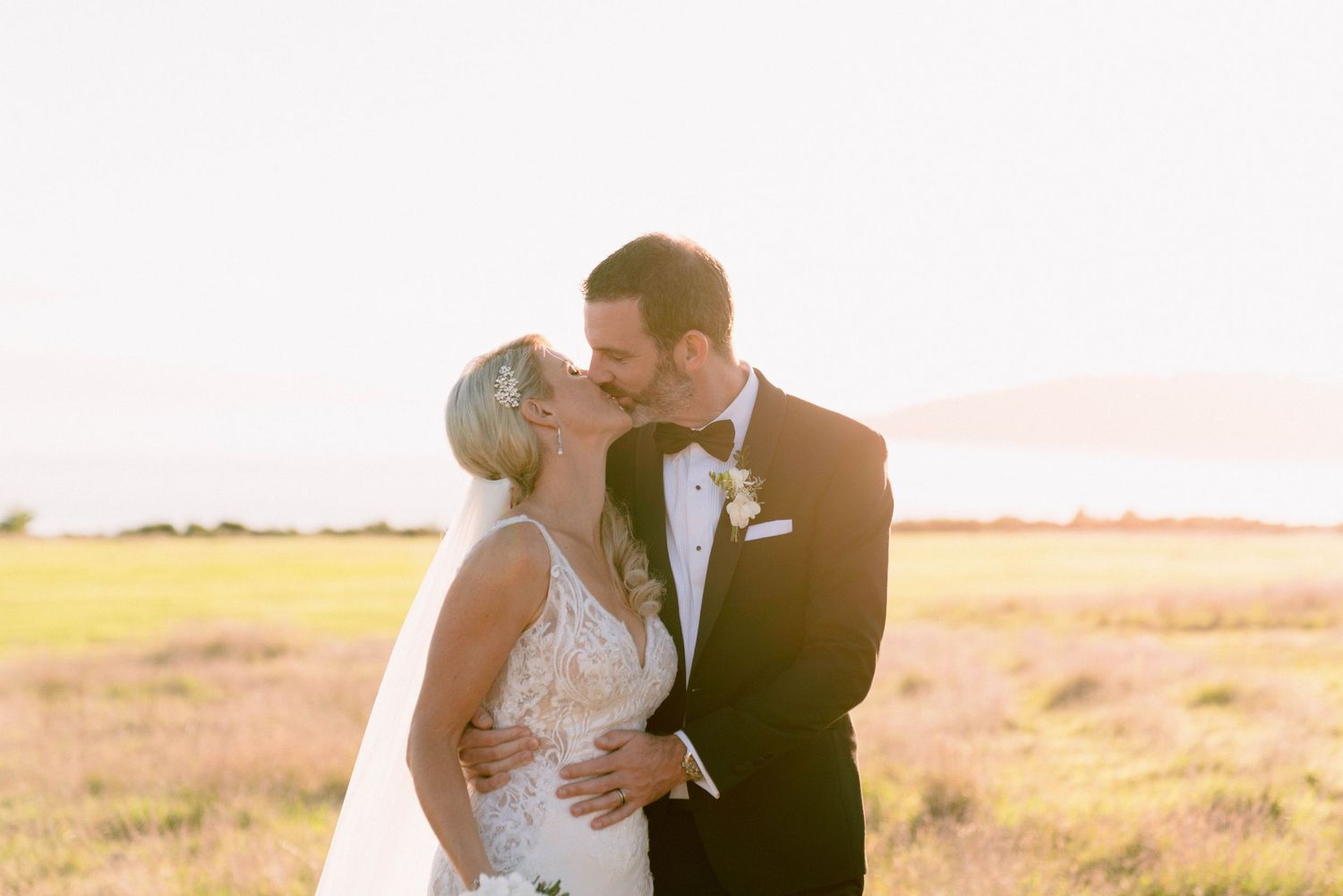 A couple kisses at sunset in a golden field while wearing formal wedding attire.