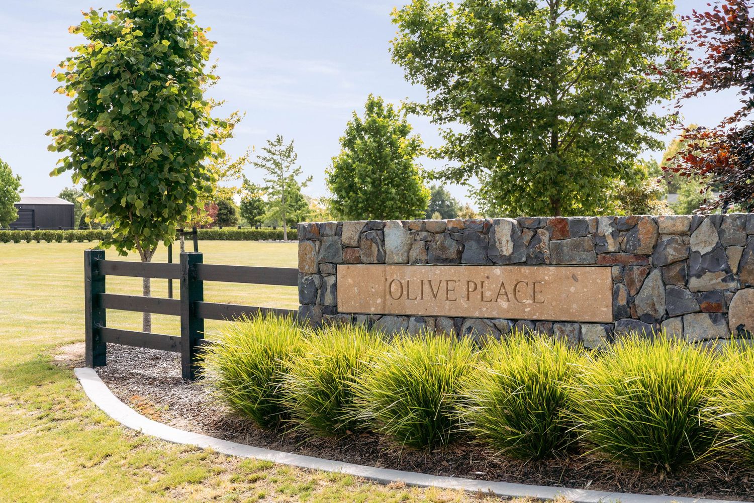 Black fence and stone wall entrance sign reading 'Olive Place' with landscaped grasses.