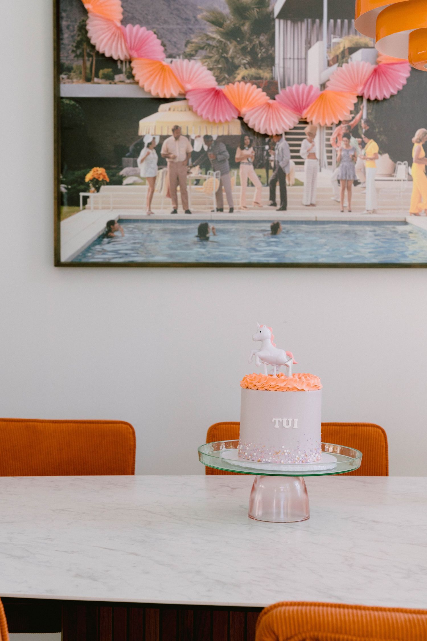 A soft pink cake with a unicorn topper sits on a glass stand beneath vintage pool party artwork with coral decorations.